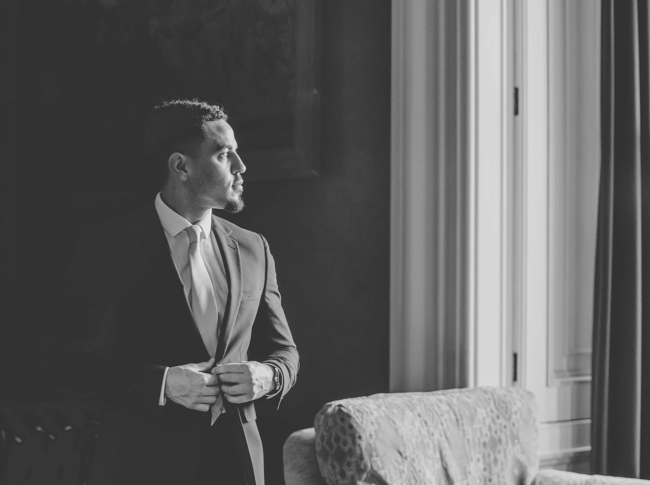 A man in a suit stands indoors, looking out a window with curtains, illuminated by natural light.