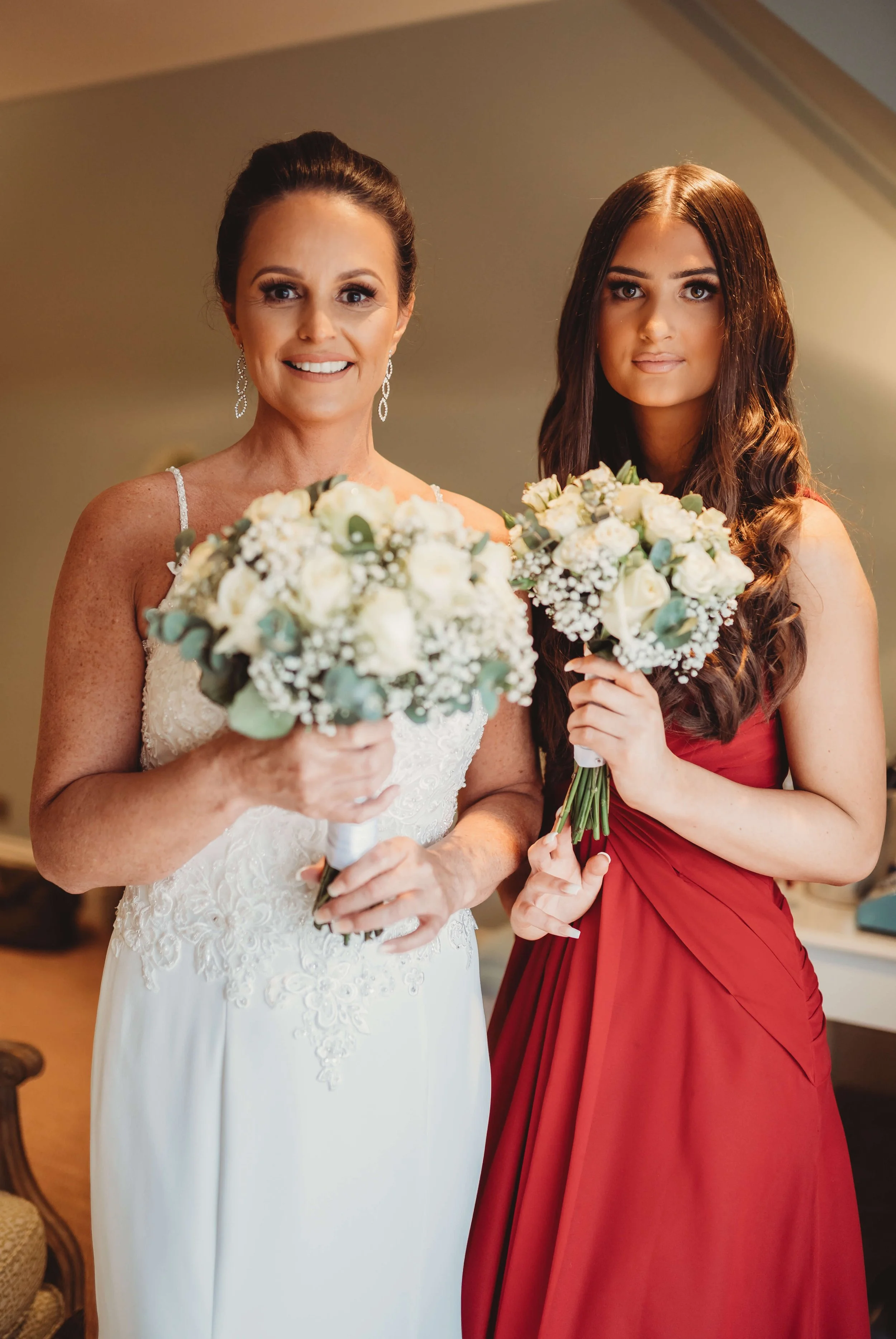 A bride in a white wedding dress and a bridesmaid in a red dress holding bouquets of white flowers, standing together indoors.