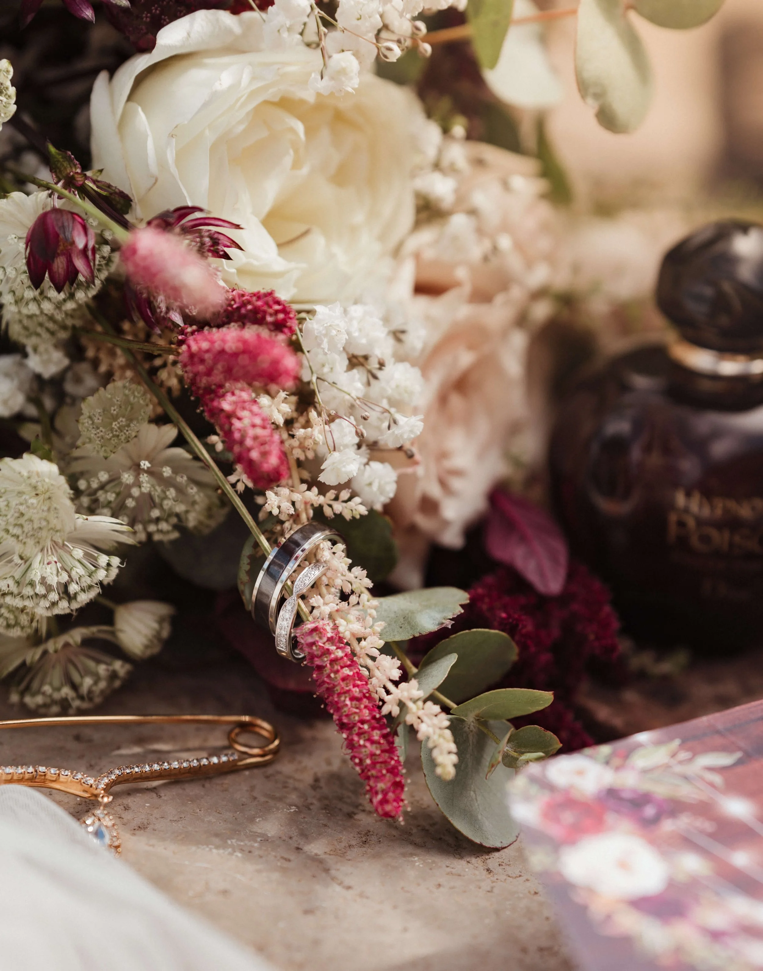 Close-up of wedding rings resting on a bouquet of flowers, including white roses and pink veronica, with a bottle of perfume and a jewelry piece nearby.