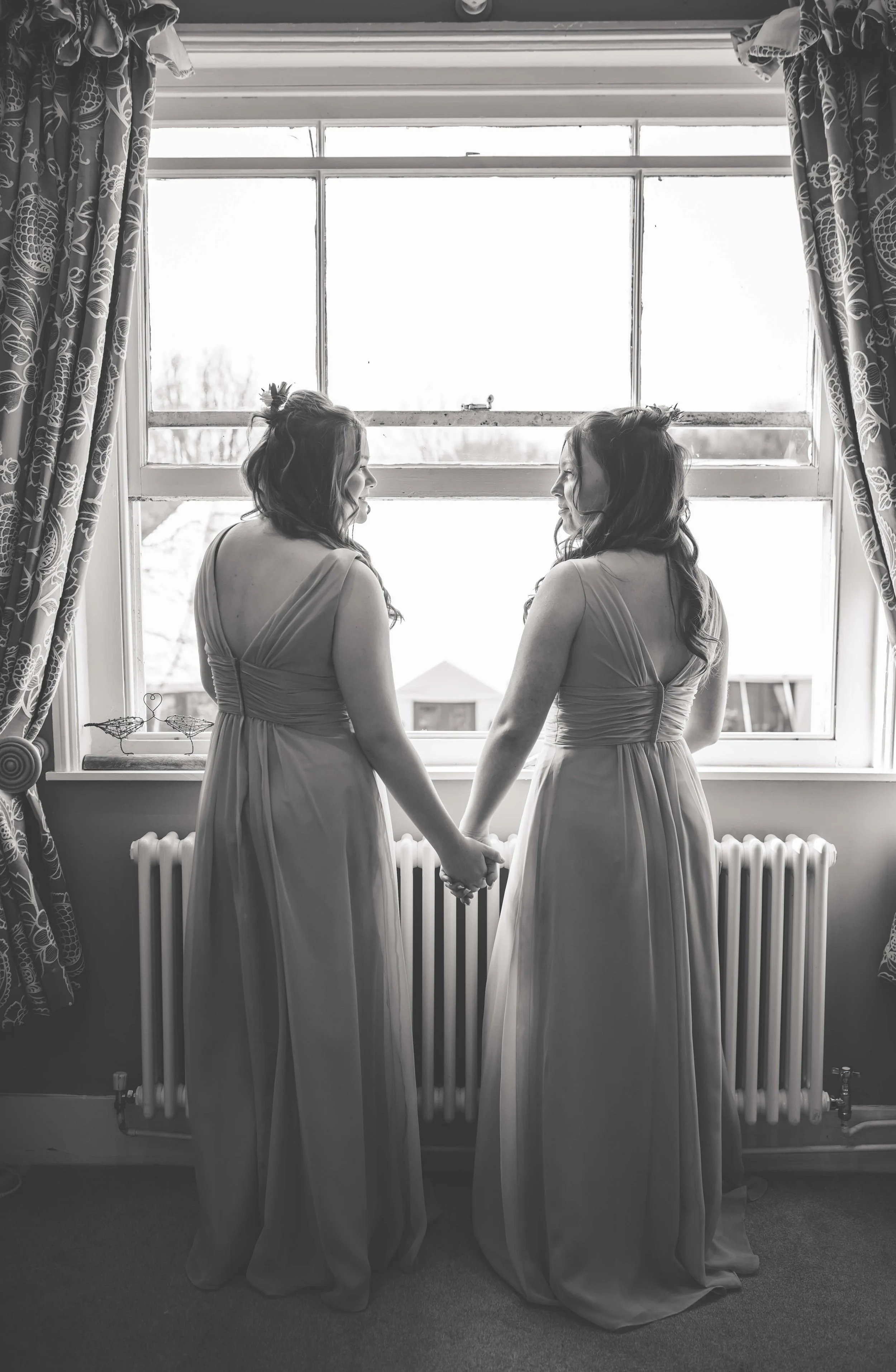 Two women in long dresses holding hands and looking at each other in front of a window, with curtains on either side.