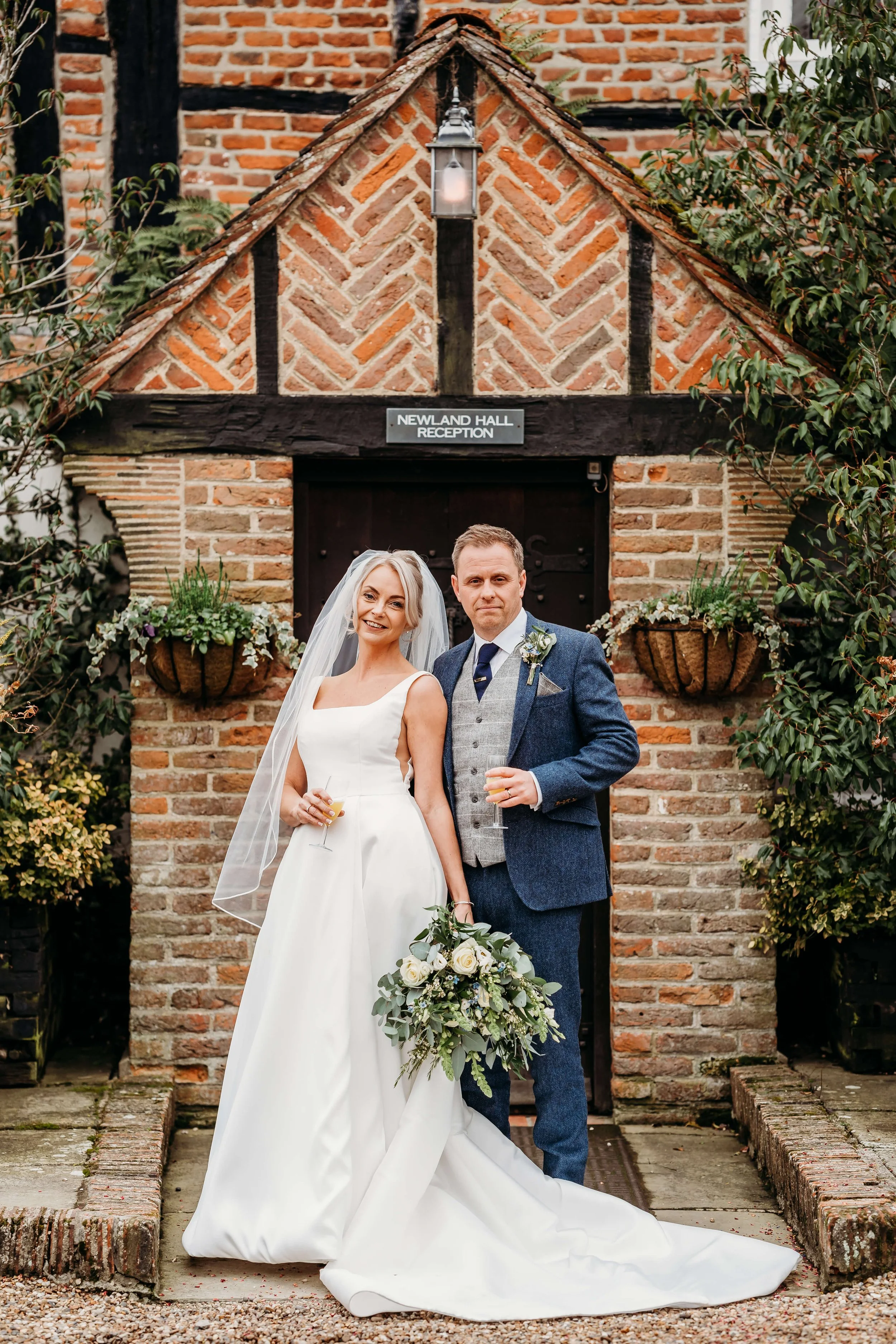 A bride and groom standing together outdoors in front of a brick building labeled 'Newland Hall Reception,' dressed in wedding attire, holding glasses and a bouquet, with greenery and hanging potted plants around them.