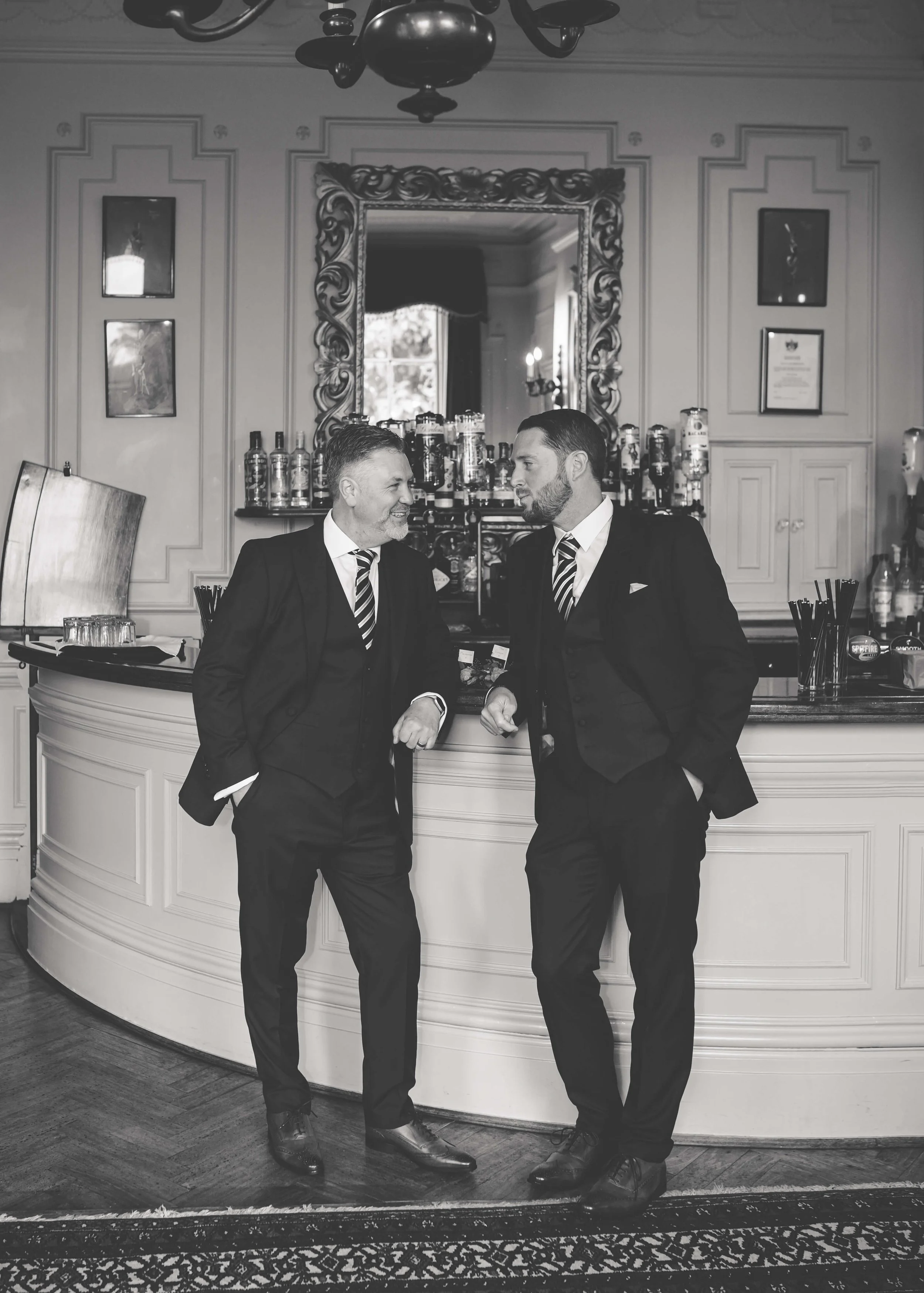 Two men in suits and striped ties standing at a bar, smiling and talking, in a room with elegant decor, a large ornate mirror, and various bottles of alcohol on the shelves behind them.