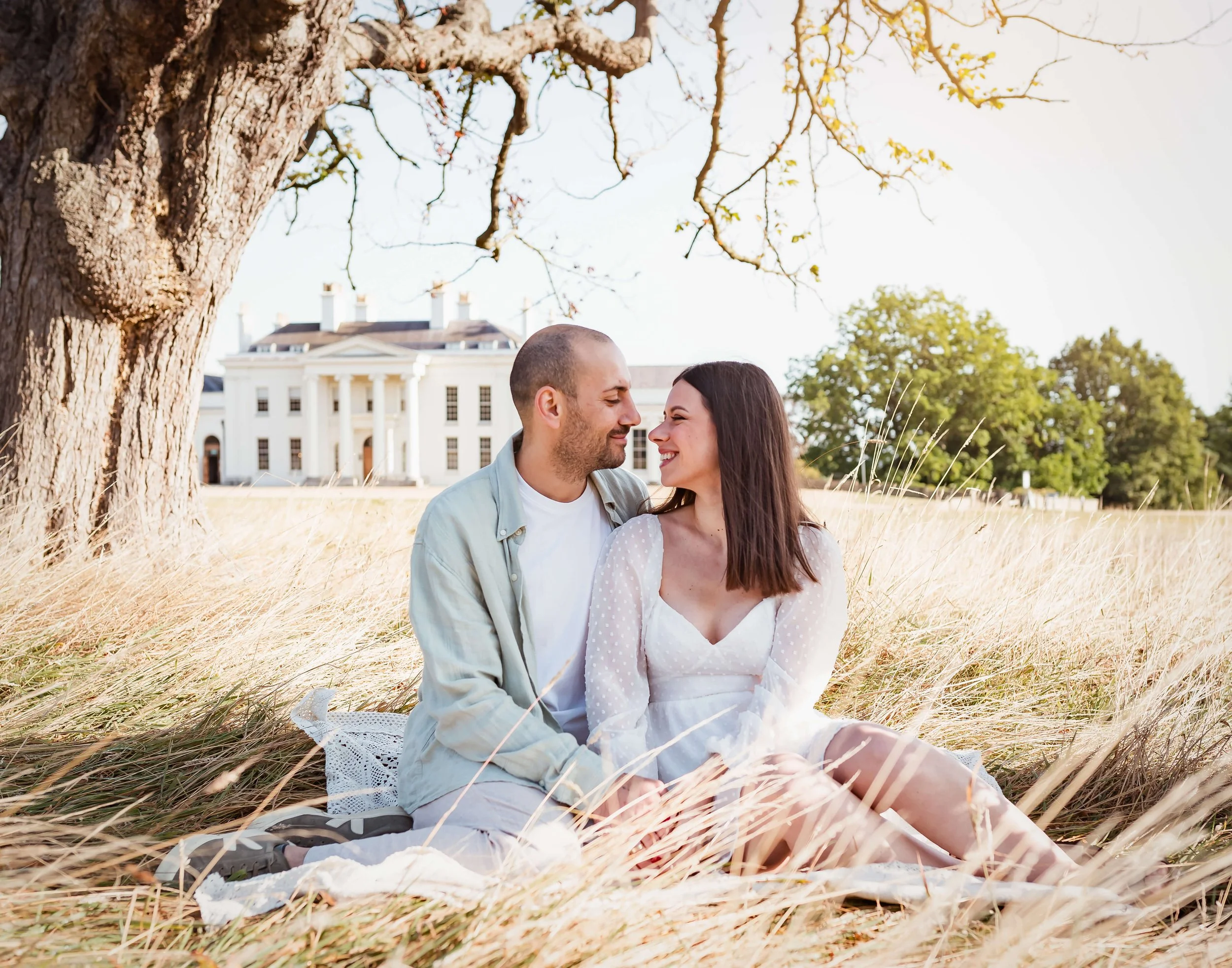 A couple sitting on a blanket in a field of tall grass, smiling and looking at each other with the White House in the background.