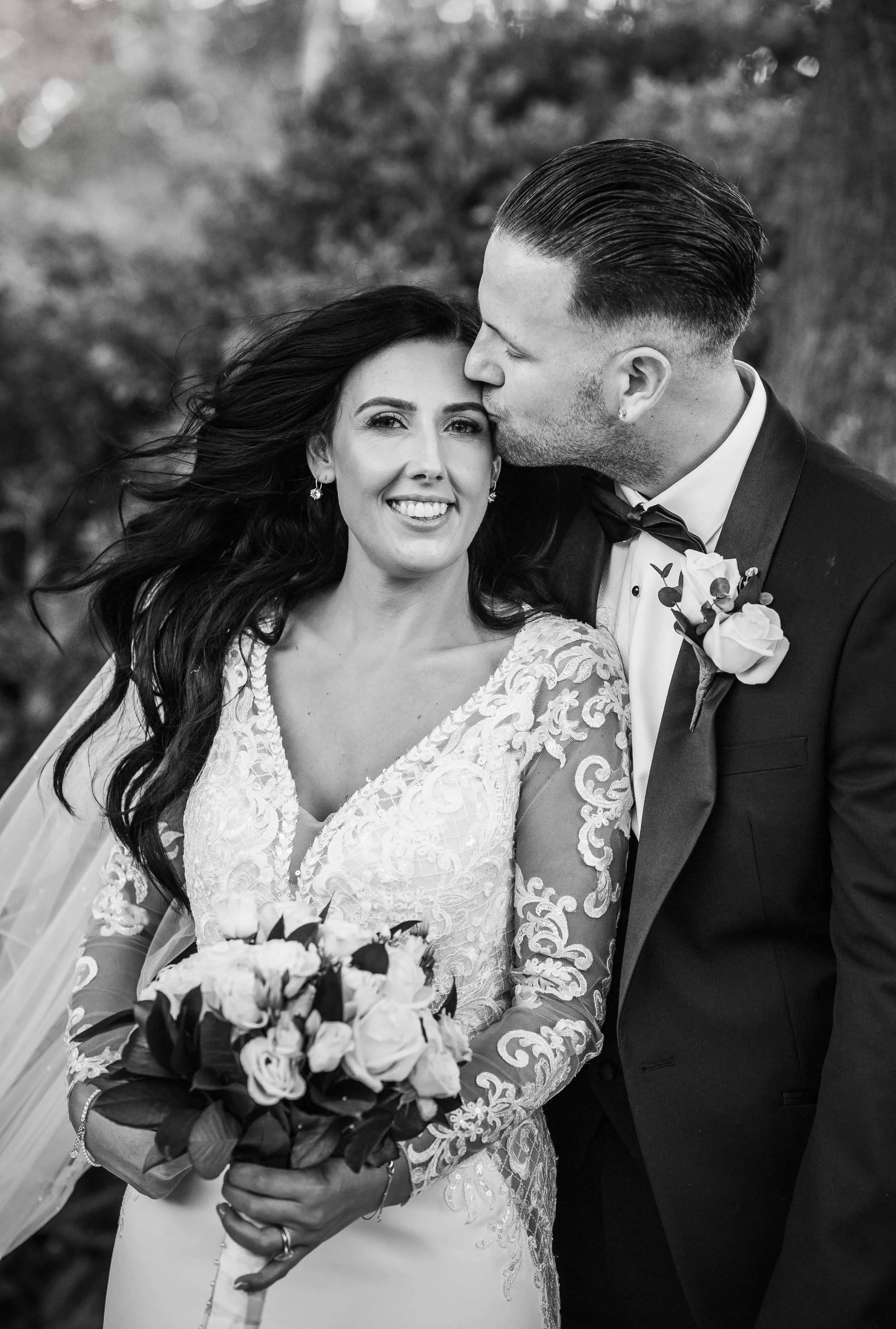 Black and white photo of a bride and groom on their wedding day outdoors. The groom is kissing the bride on the forehead, and she is holding a bouquet of flowers and smiling.