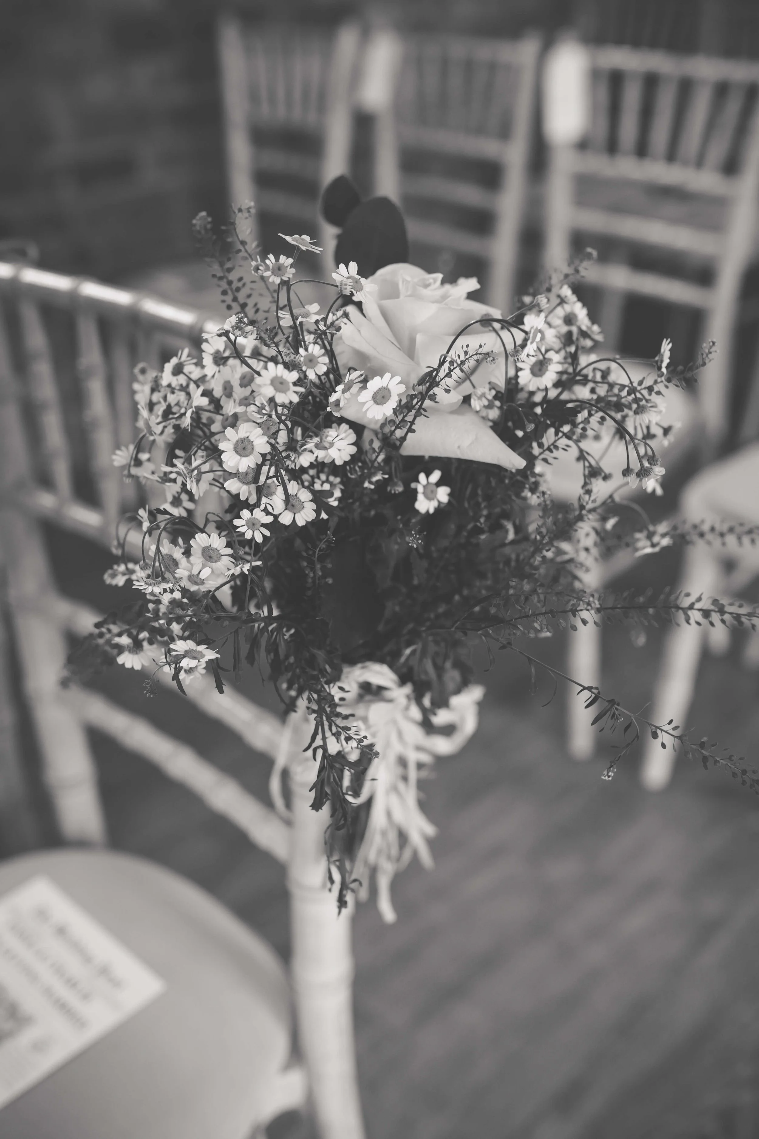 A black-and-white photo of a small flower arrangement with a white rose and tiny daisies, attached to a chair at an event or gathering.