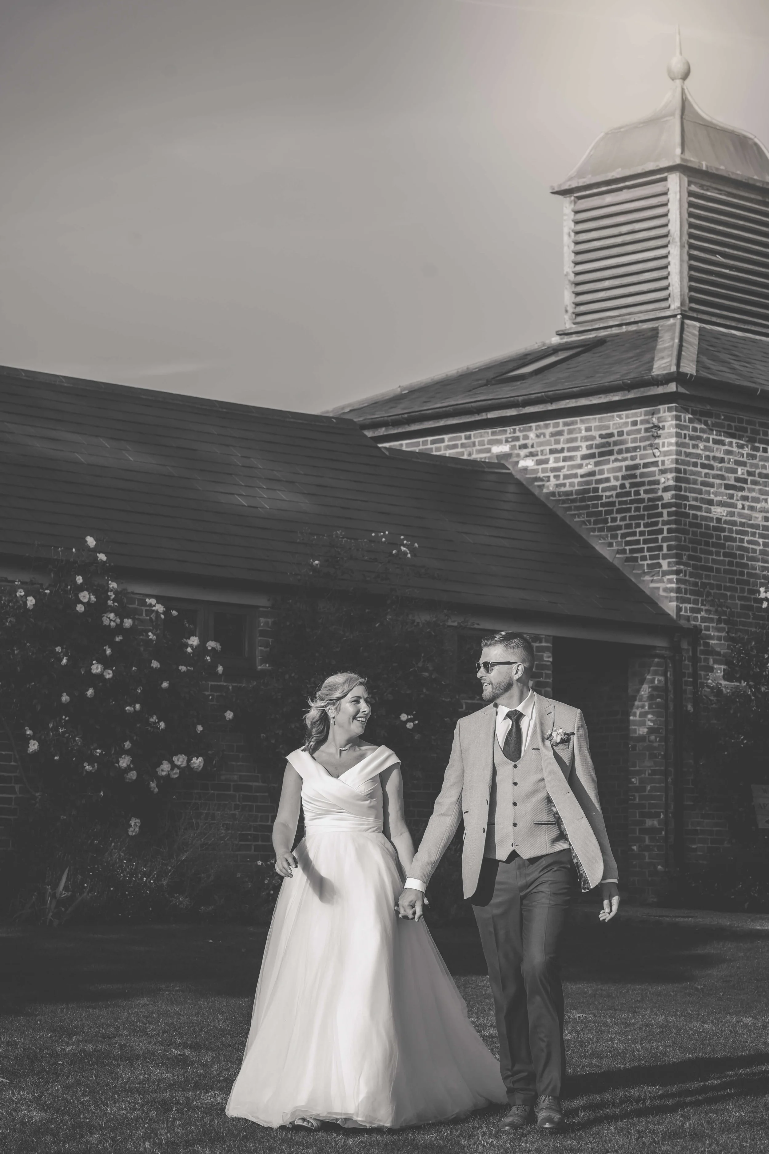Black and white photo of a smiling newlywed couple walking hand in hand outside a brick building, possibly a church, during daytime.