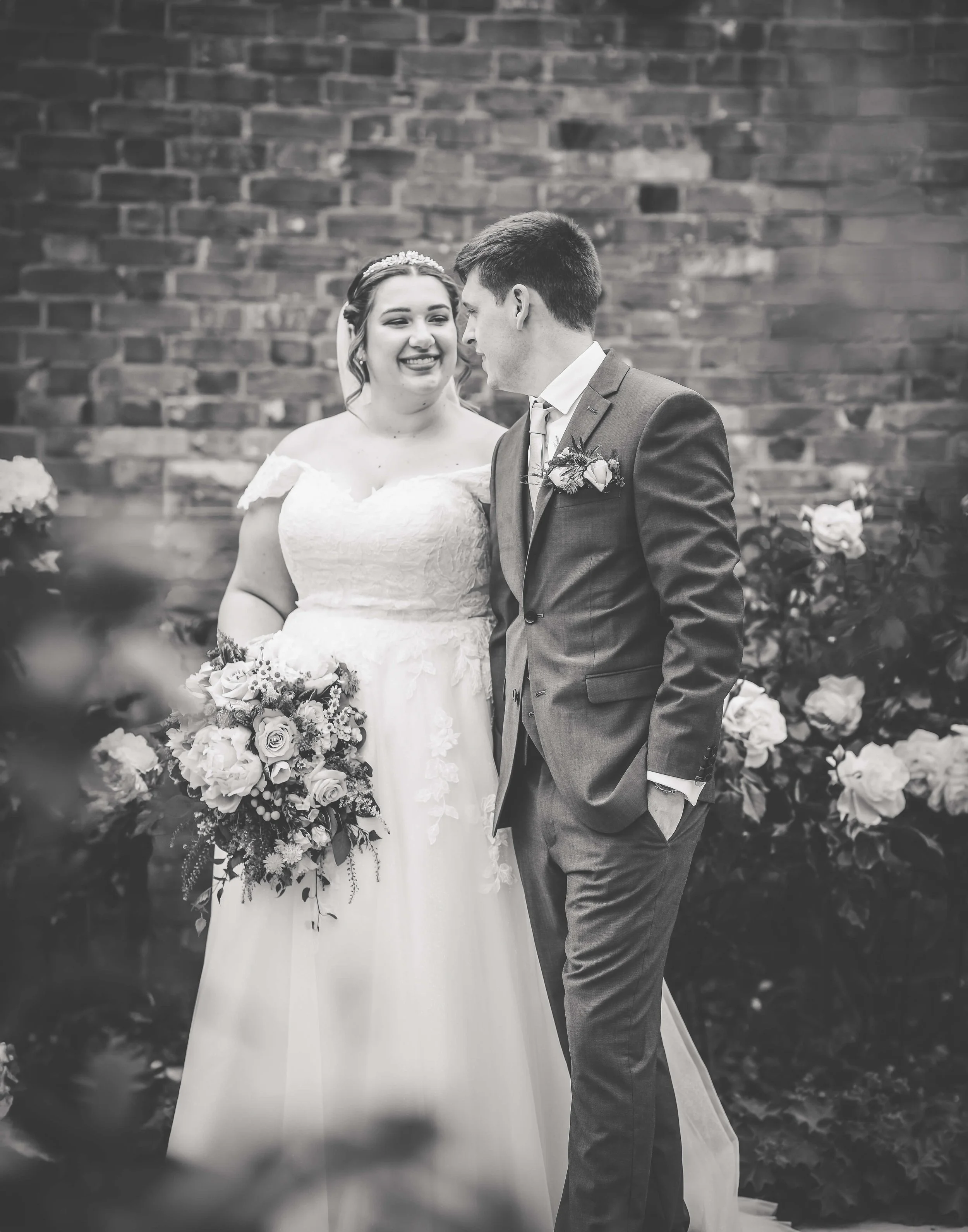 A bride and groom on their wedding day, smiling and looking at each other, standing in front of a brick wall with flowers in the background.
