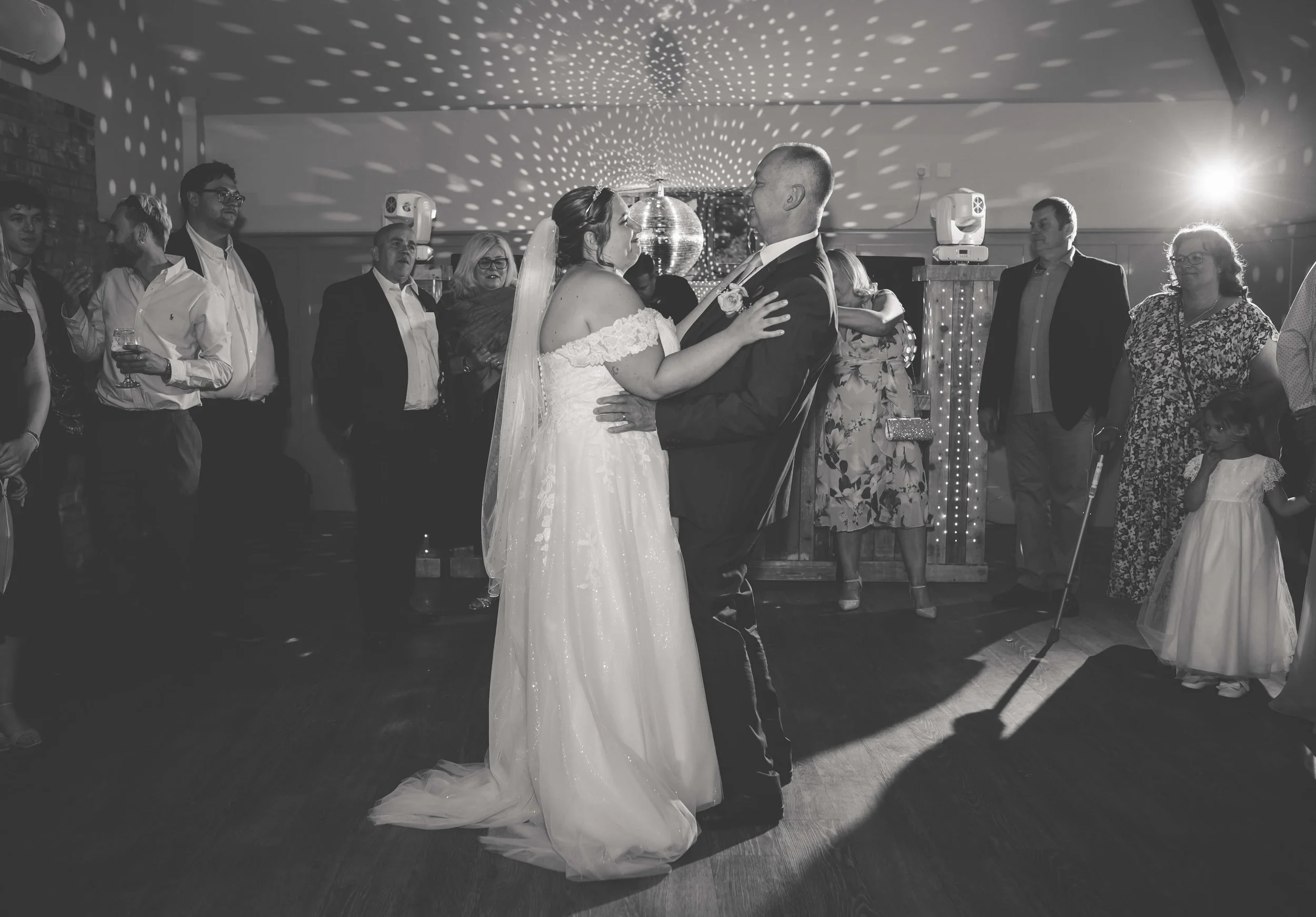 A bride and groom are dancing together at their wedding reception surrounded by guests, with a disco ball and light patterns on the ceiling.