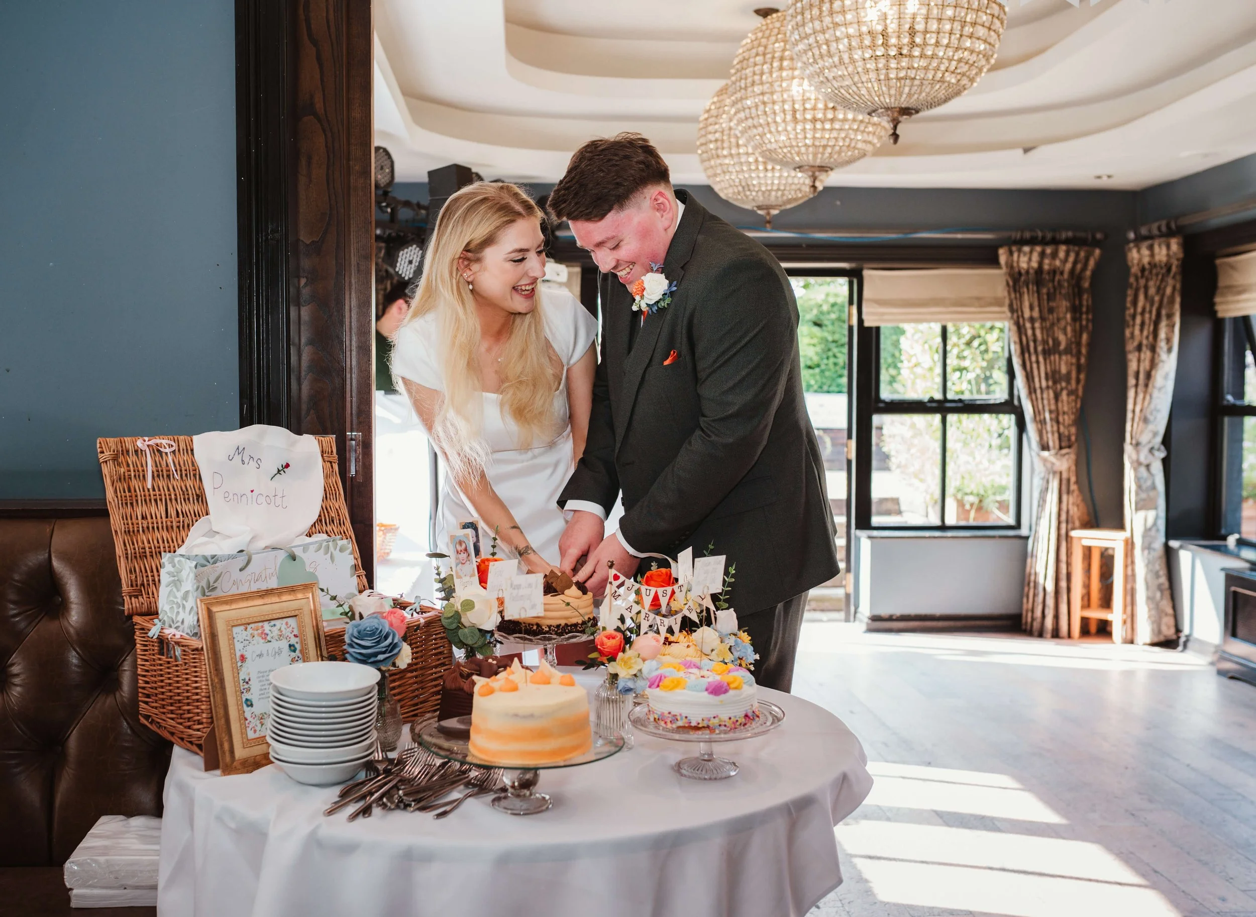 A newlywed couple is cutting their wedding cake at a celebration. The bride has long blonde hair and is wearing a white dress, and the groom is in a dark suit. There are multiple decorated cakes on the table, along with gift bags and framed photos.