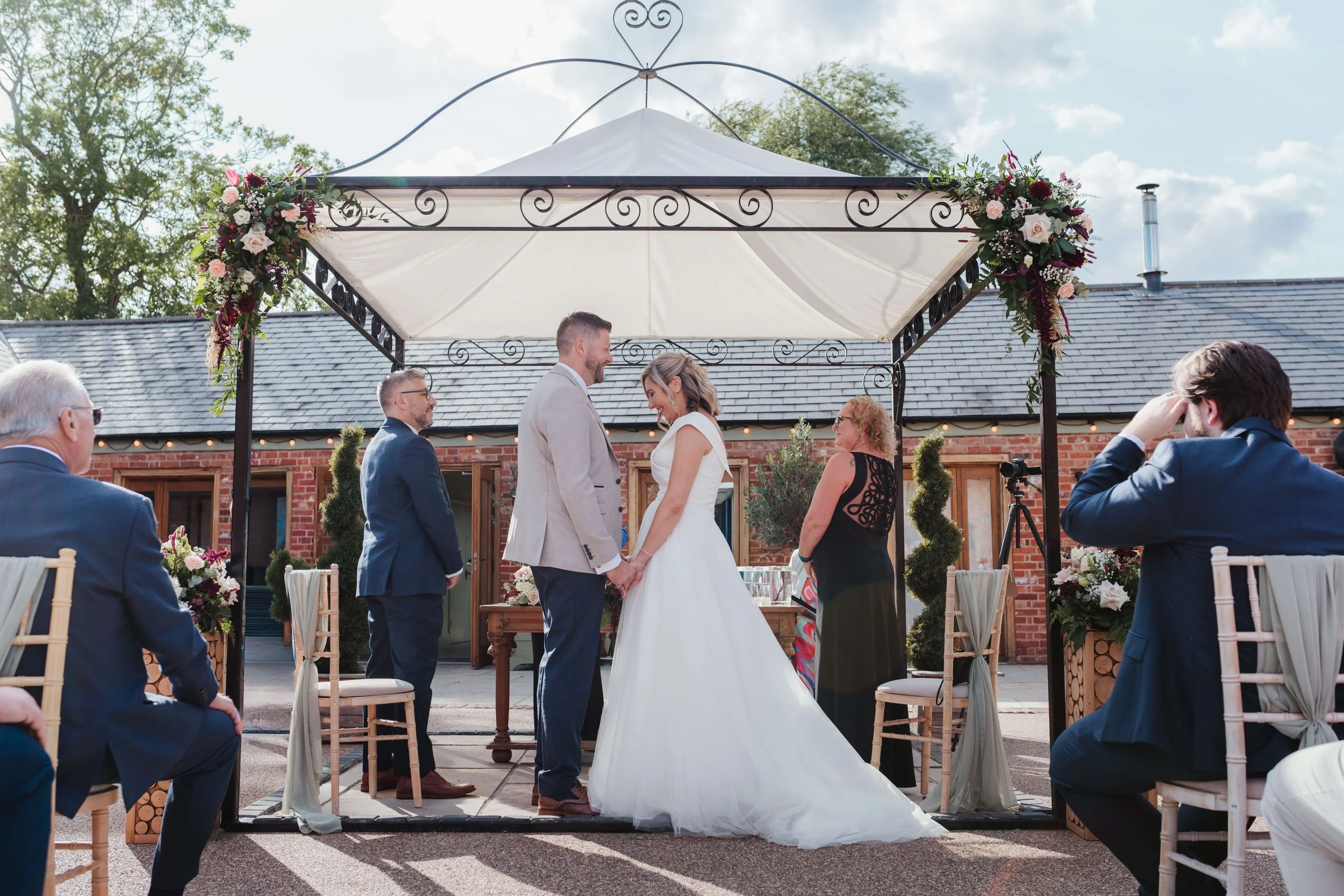 A wedding ceremony outdoors under a white canopy with floral decorations. The bride and groom are holding hands, facing each other, with the officiant and guests seated around them.