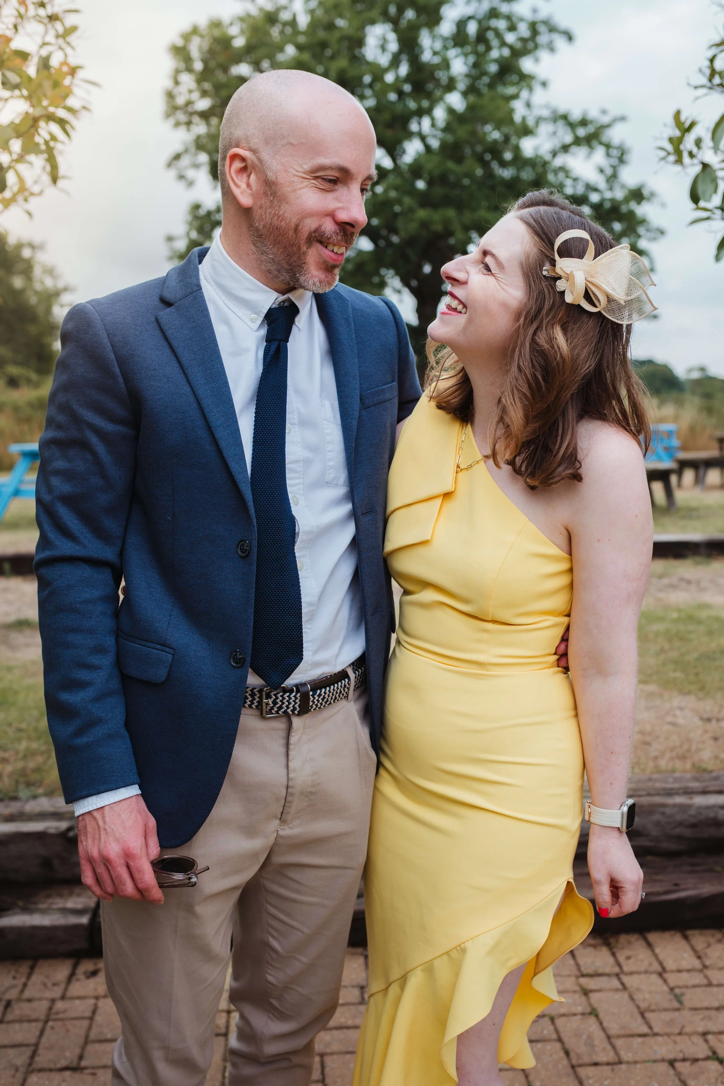 A man in a suit and a woman in a yellow dress are smiling and looking at each other outdoors.