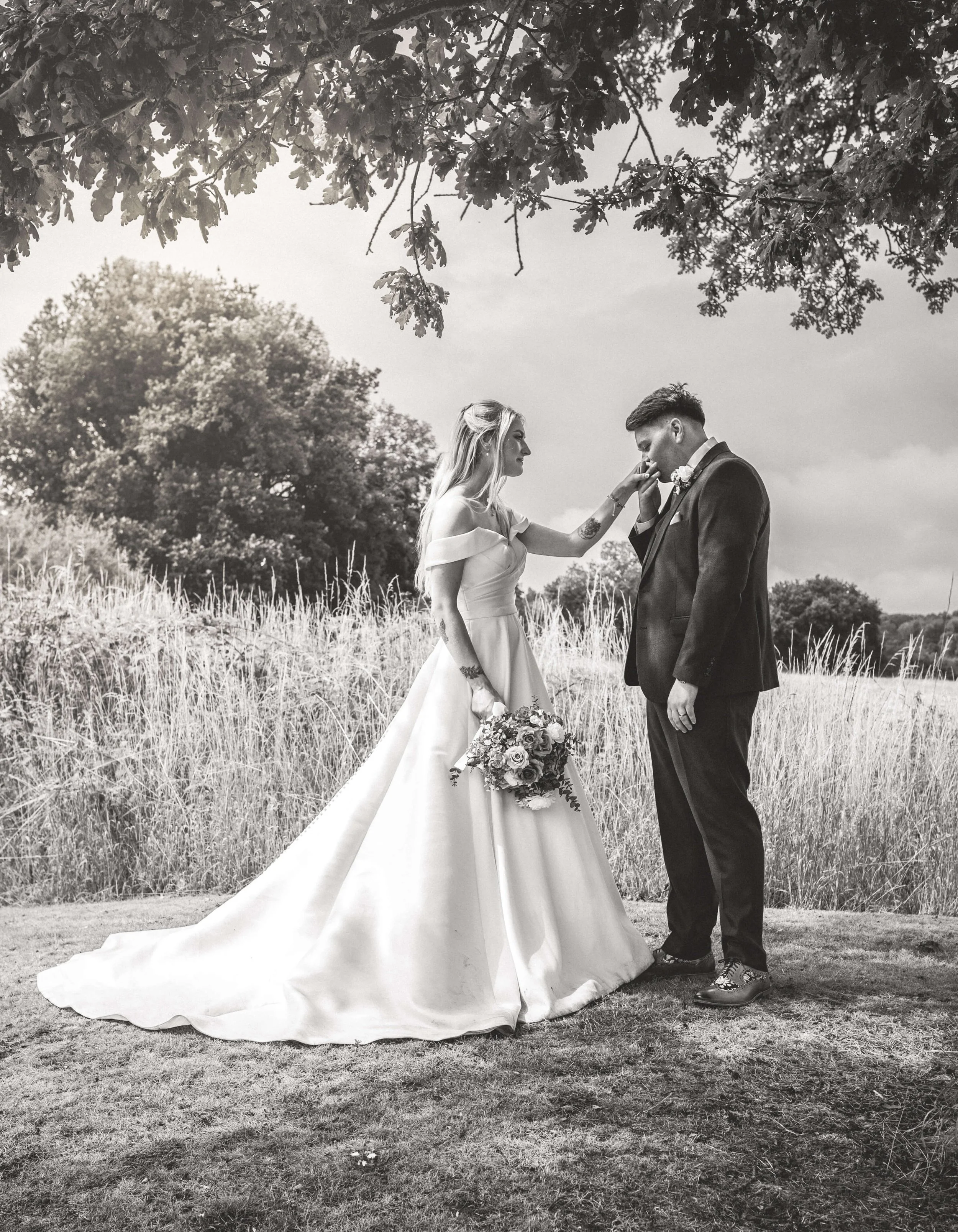 Black and white photo of a bride and groom outdoors, with the bride gently touching the groom's face. The bride is wearing an off-the-shoulder wedding dress and holding a bouquet, while the groom is in a suit. They are standing on grass in a natural 