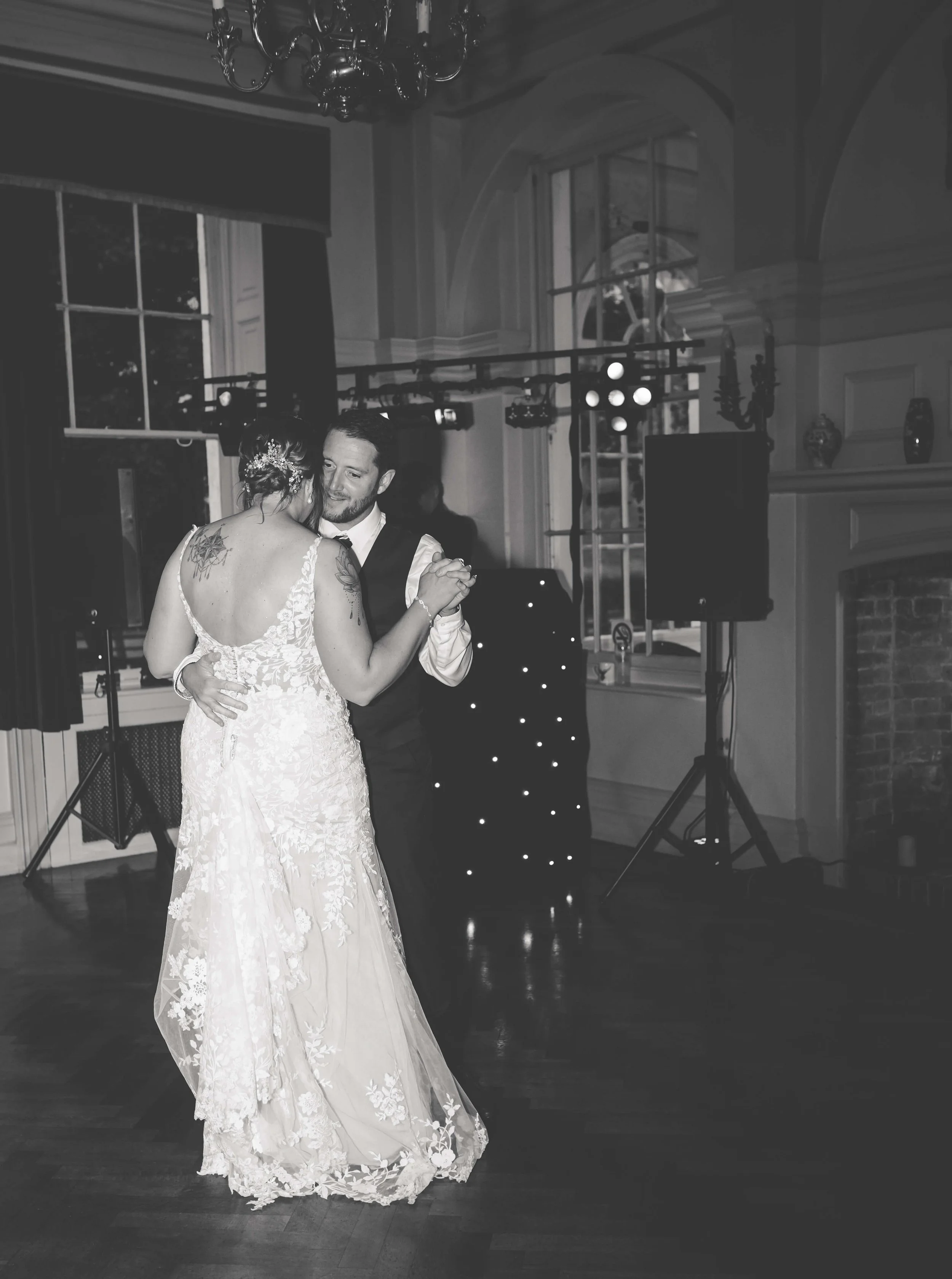 A bride and groom sharing a dance at their wedding reception in a decorated indoor venue, with lighting equipment and large windows in the background.
