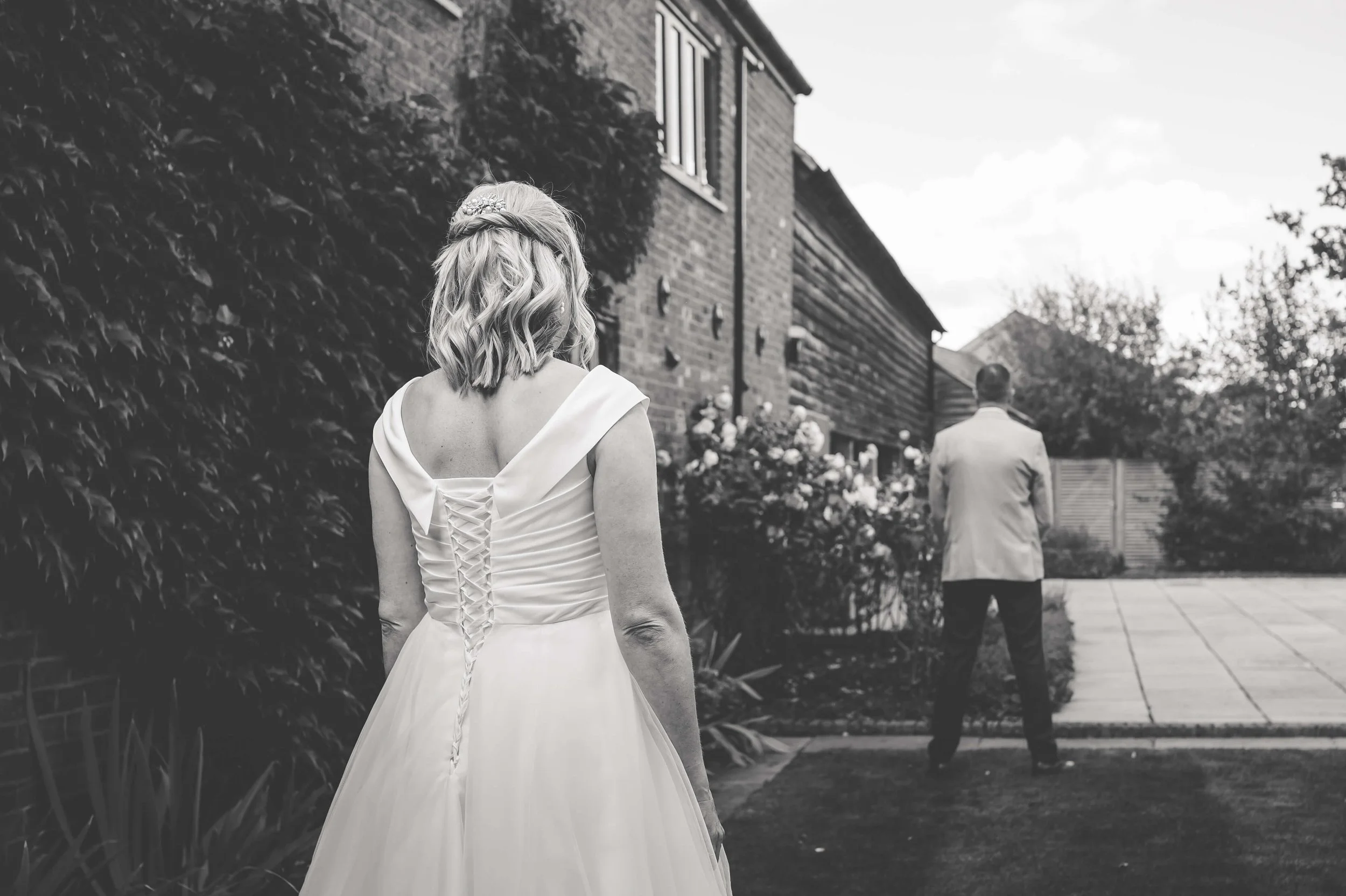 A bride in a wedding dress standing outdoors, looking at a man at a distance, near a house with flowers and trees in the background.