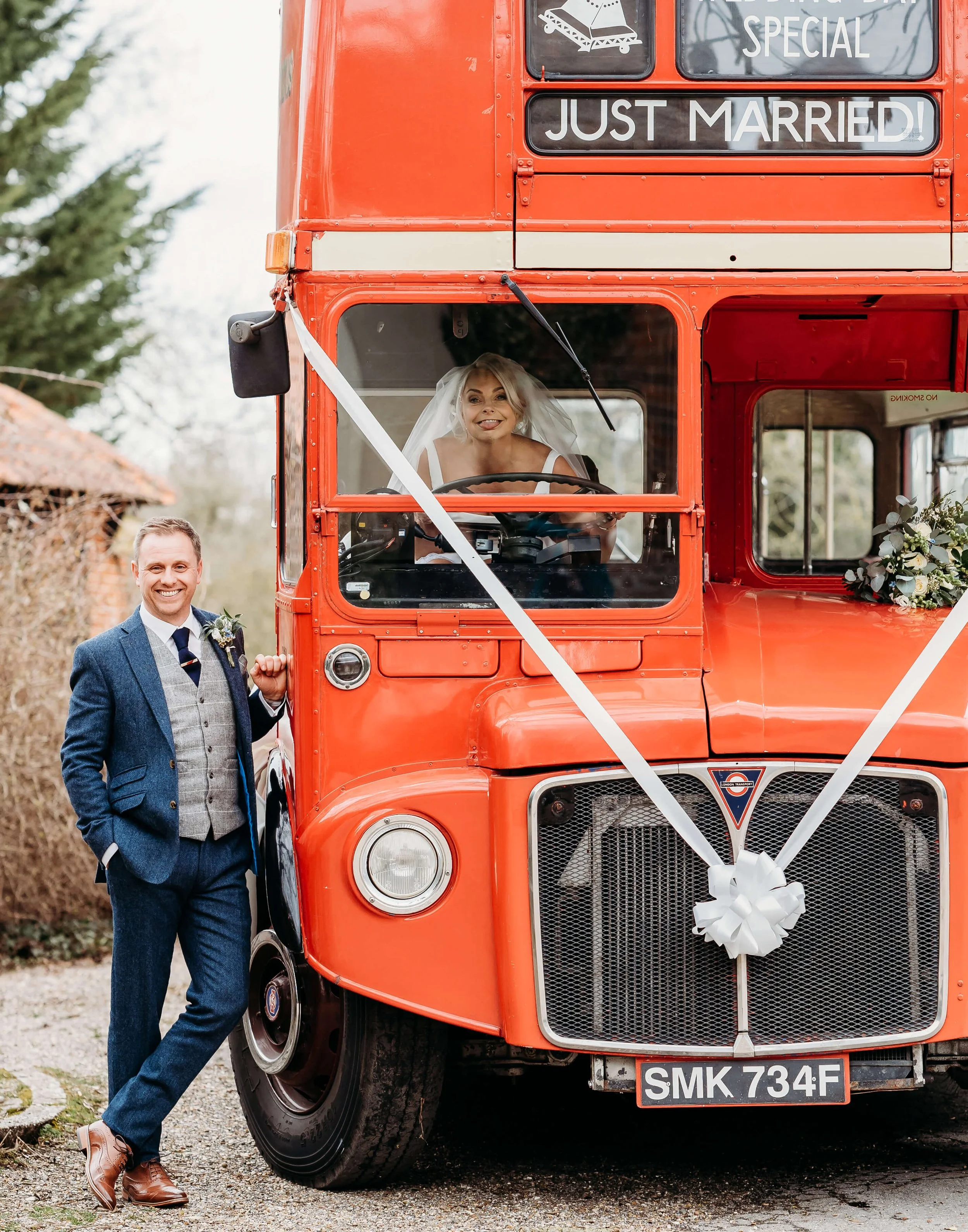 A smiling woman in a wedding dress and veil is sitting in the driver's seat of a red double-decker bus decorated with a white ribbon and bow, marked with 'Just Married!'. A man in a blue suit, gray vest, and tie is standing next to the bus, holding t