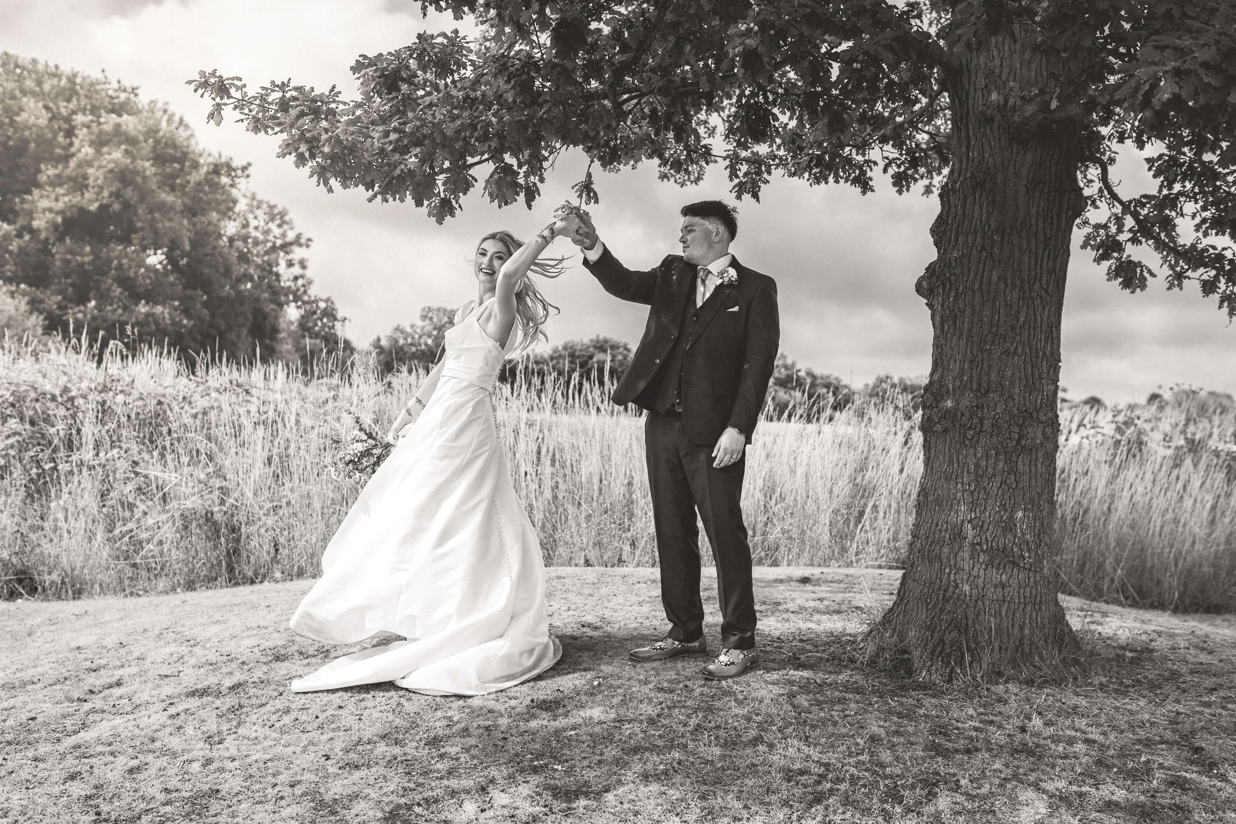 A bride and groom dancing outdoors under a tree in a field, black and white photo, the bride is wearing a white wedding gown, and the groom is in a suit, holding hands and smiling.