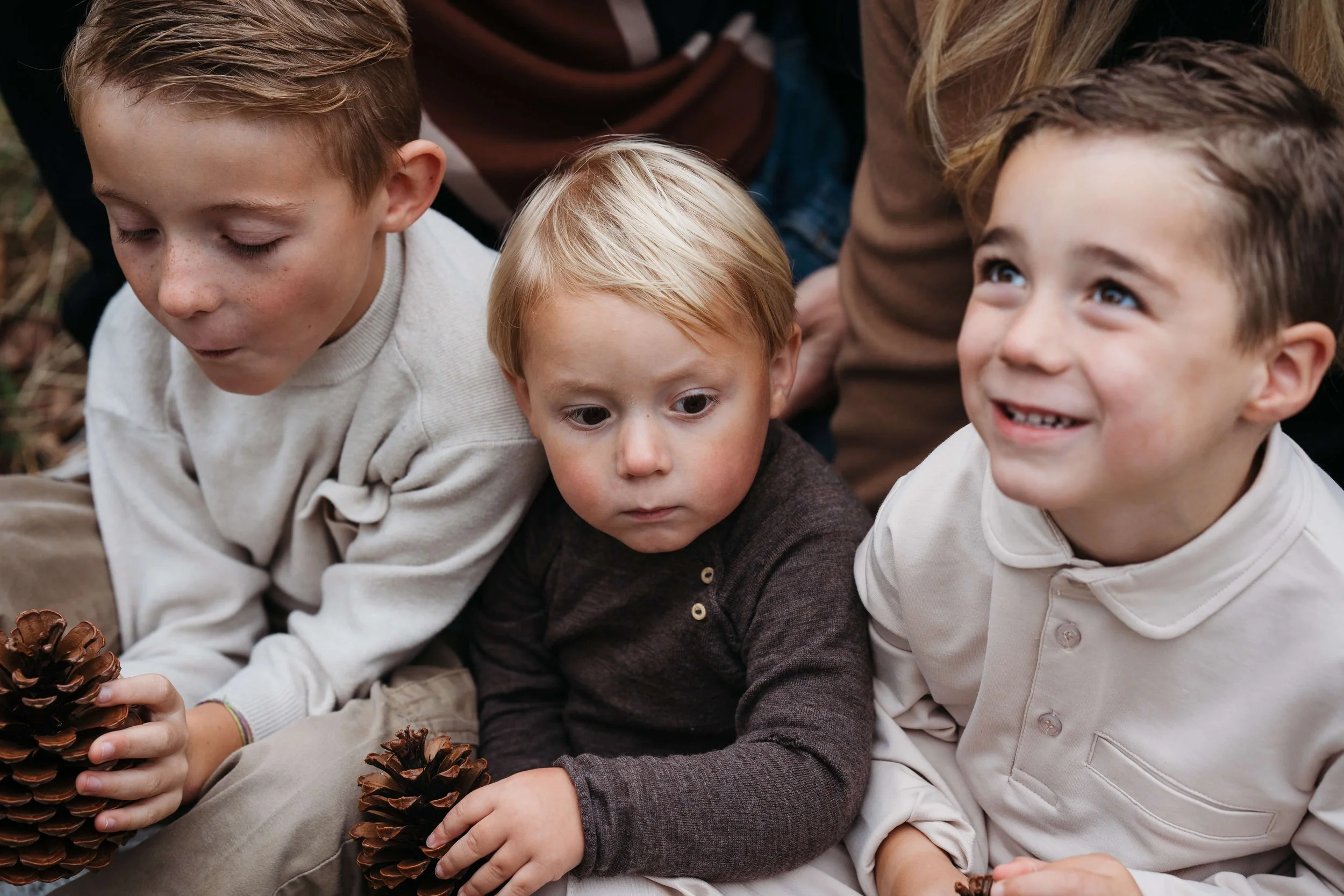 Three young boys sitting closely together outdoors, holding pinecones, with a woman partially visible behind them, smiling and engaging with the children.