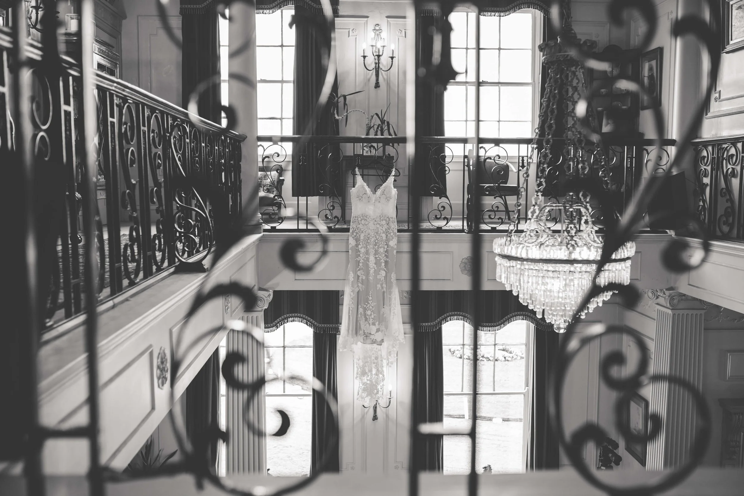A wedding dress hanging in a grand, well-lit room with large windows, ornate railings, a chandelier, and classical decor.