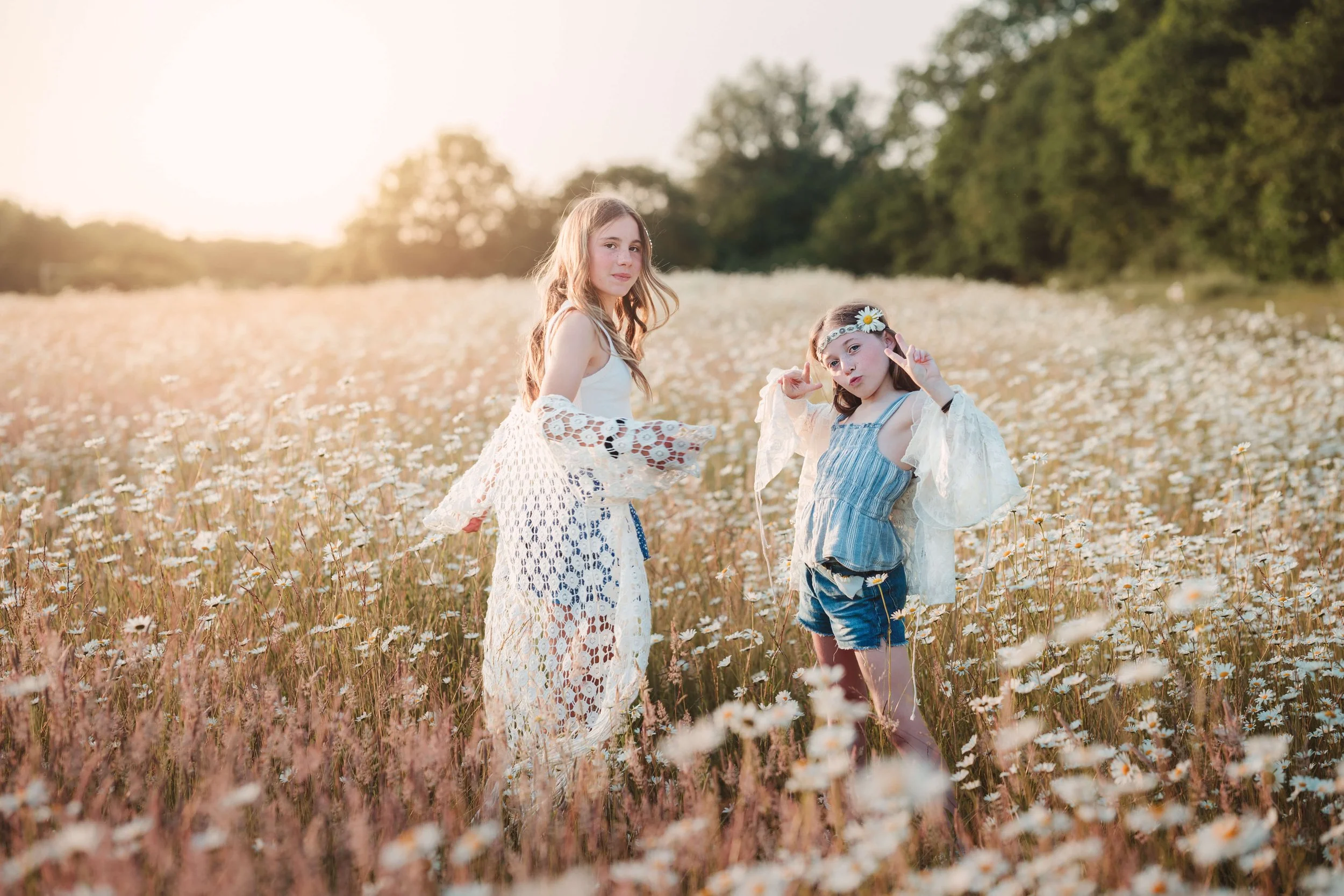 Two young girls standing in a field of daisies during sunset, dressed in summer clothes with lace accessories, making playful gestures.
