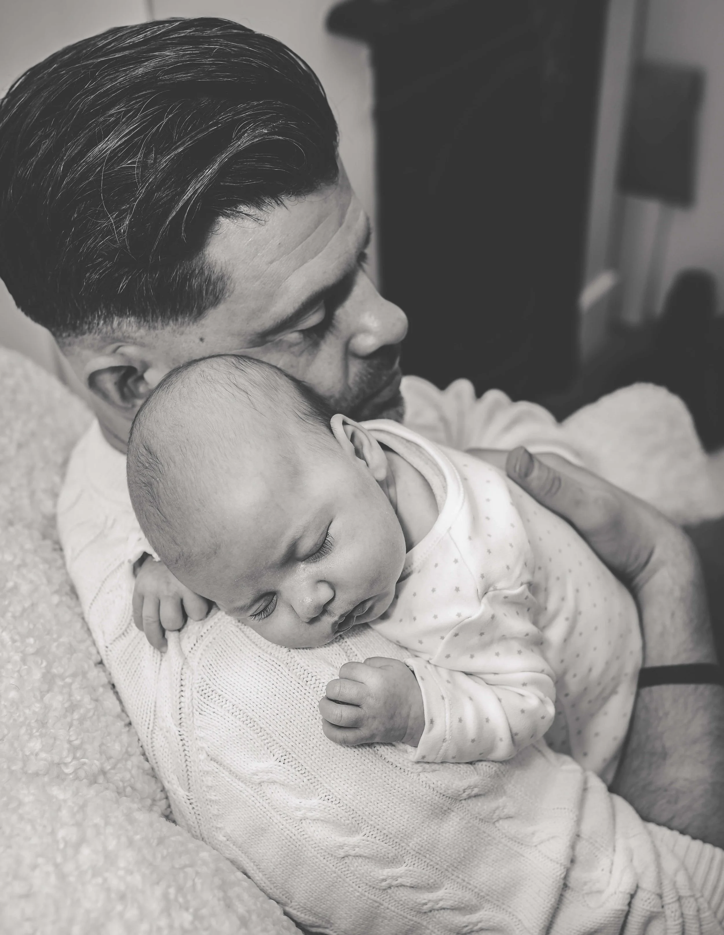 A man holding a sleeping baby close, both resting on a soft surface. The image is in black and white.