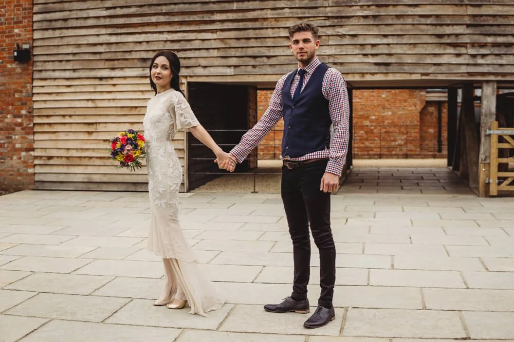A couple holding hands outdoors, with the woman in a white dress holding a colorful bouquet, and the man in a checkered shirt, vest, and dark pants, standing on a paved area in front of a wooden building.