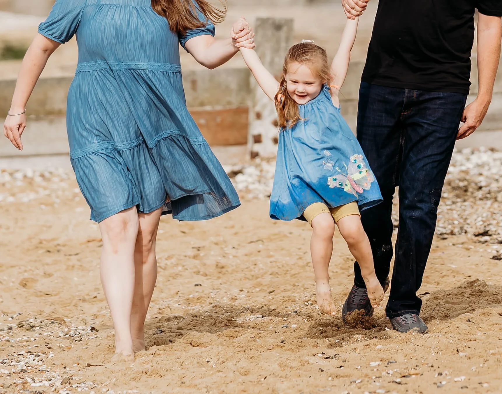 A family of three walking barefoot on a sandy beach, holding hands, and smiling. The little girl is wearing a blue dress with a butterfly embroidery, and her parents are dressed casually.