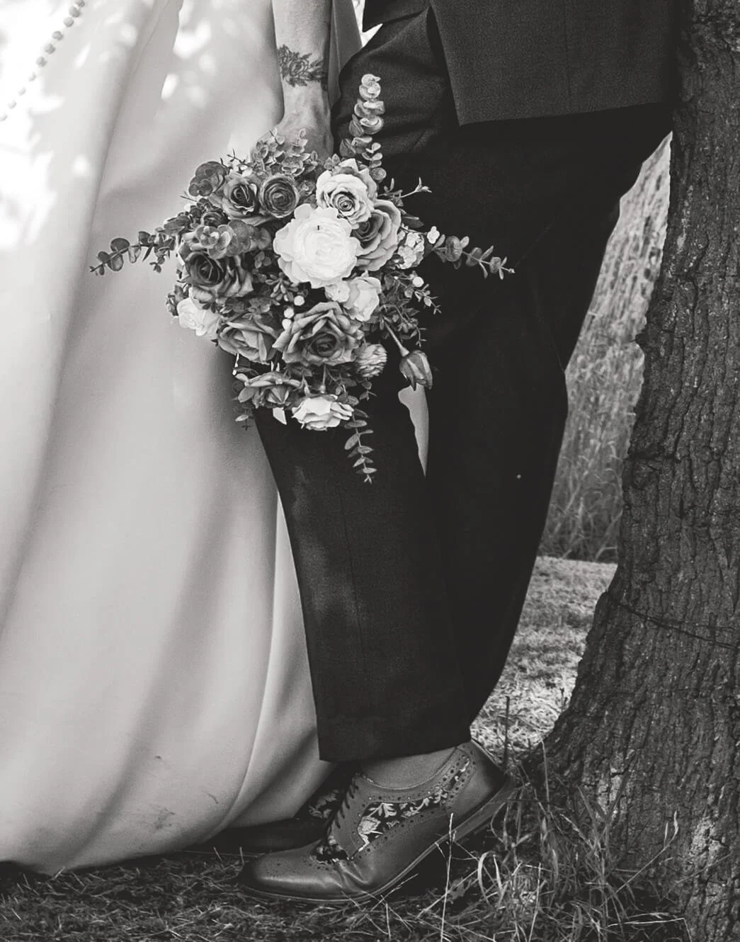 Close-up of a bride and groom standing outdoors, with the bride holding a bouquet of flowers, the groom leaning against a tree, both dressed in wedding attire, black and white photograph.