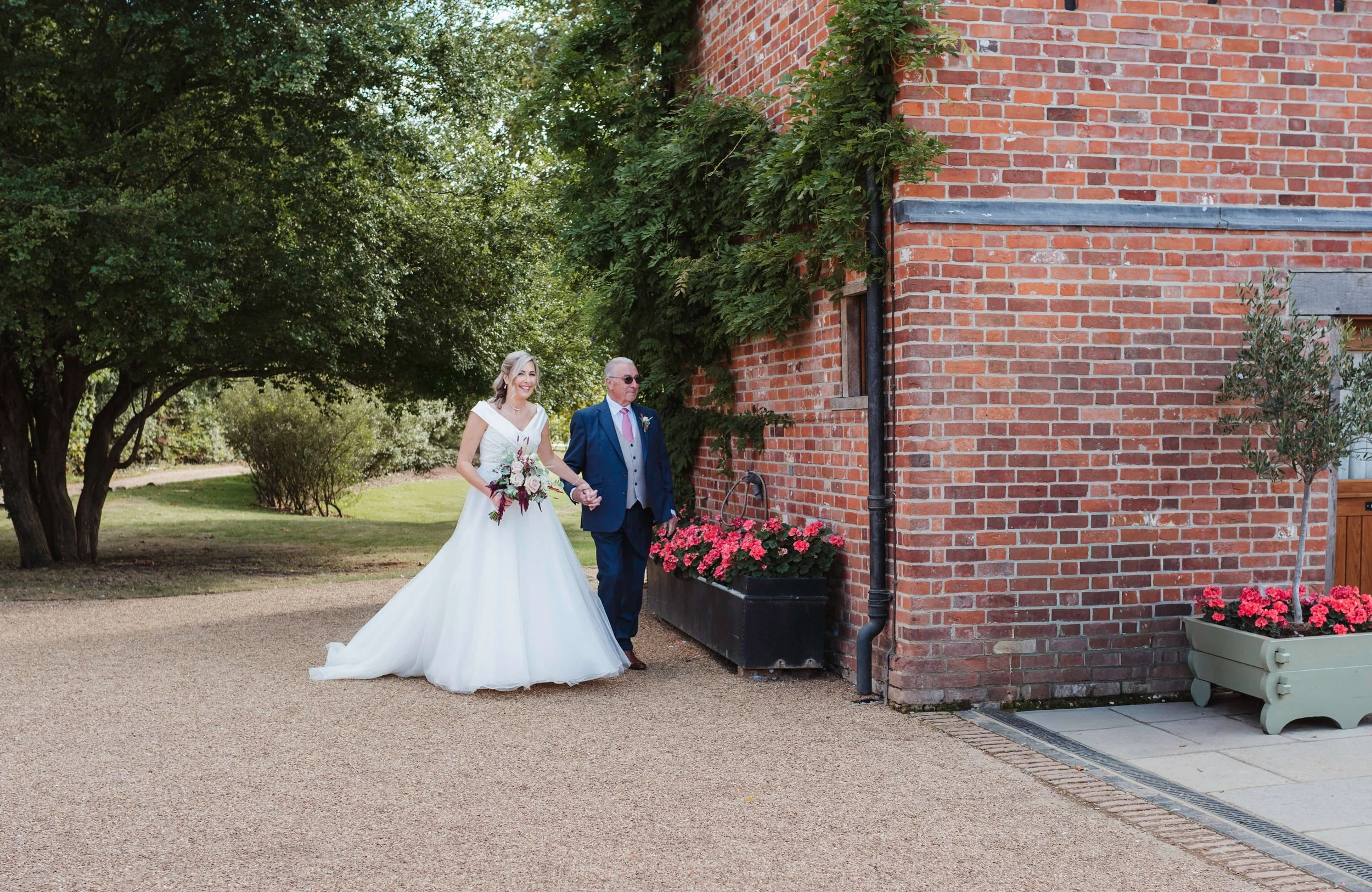 Bride in a white wedding dress holding a bouquet walking with an older man in a suit along a brick building with flowers, next to green trees.