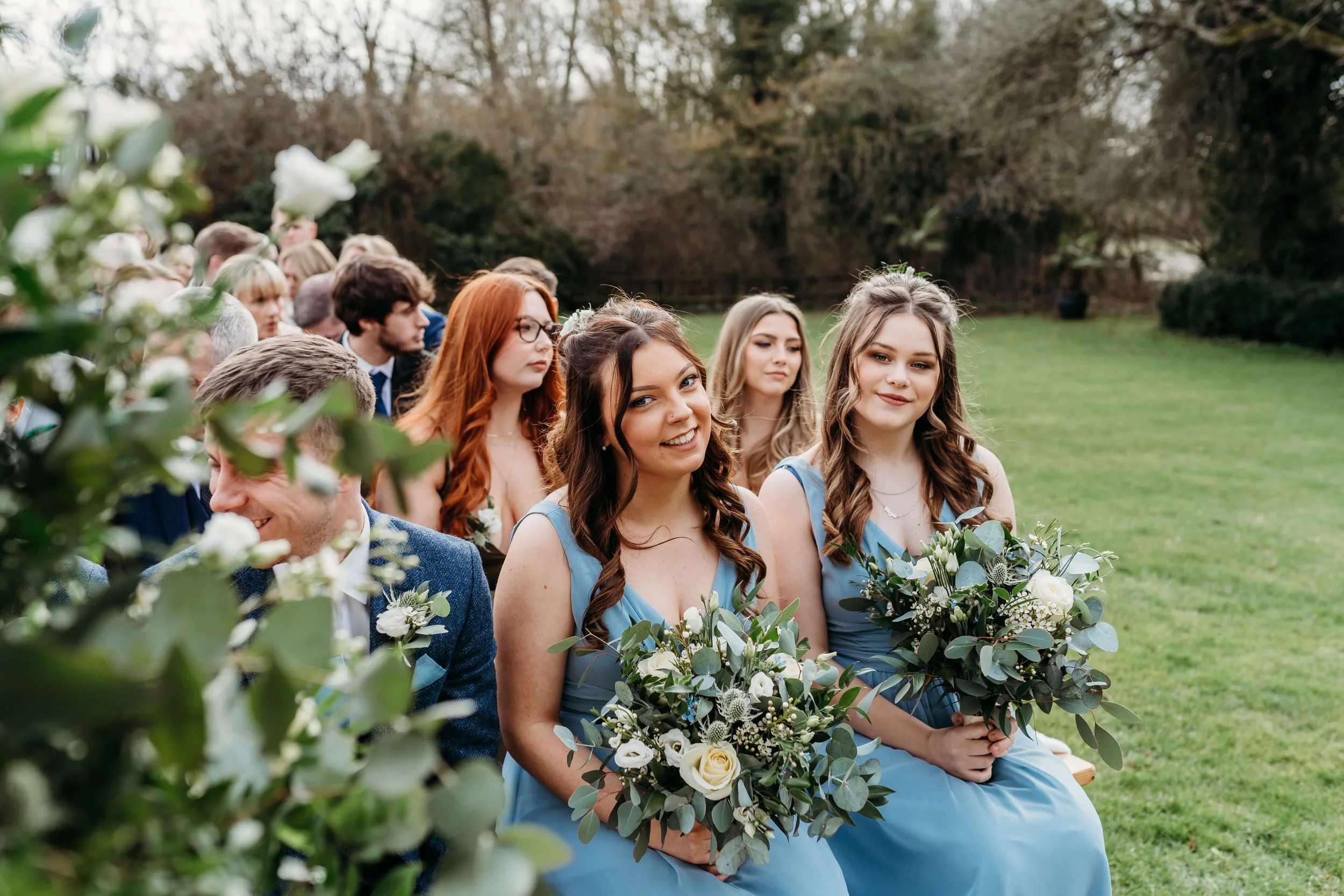 Group of bridesmaids and groomsmen sitting outdoors during wedding ceremony, wearing matching light blue dresses and suits, holding bouquets of white flowers and greenery.