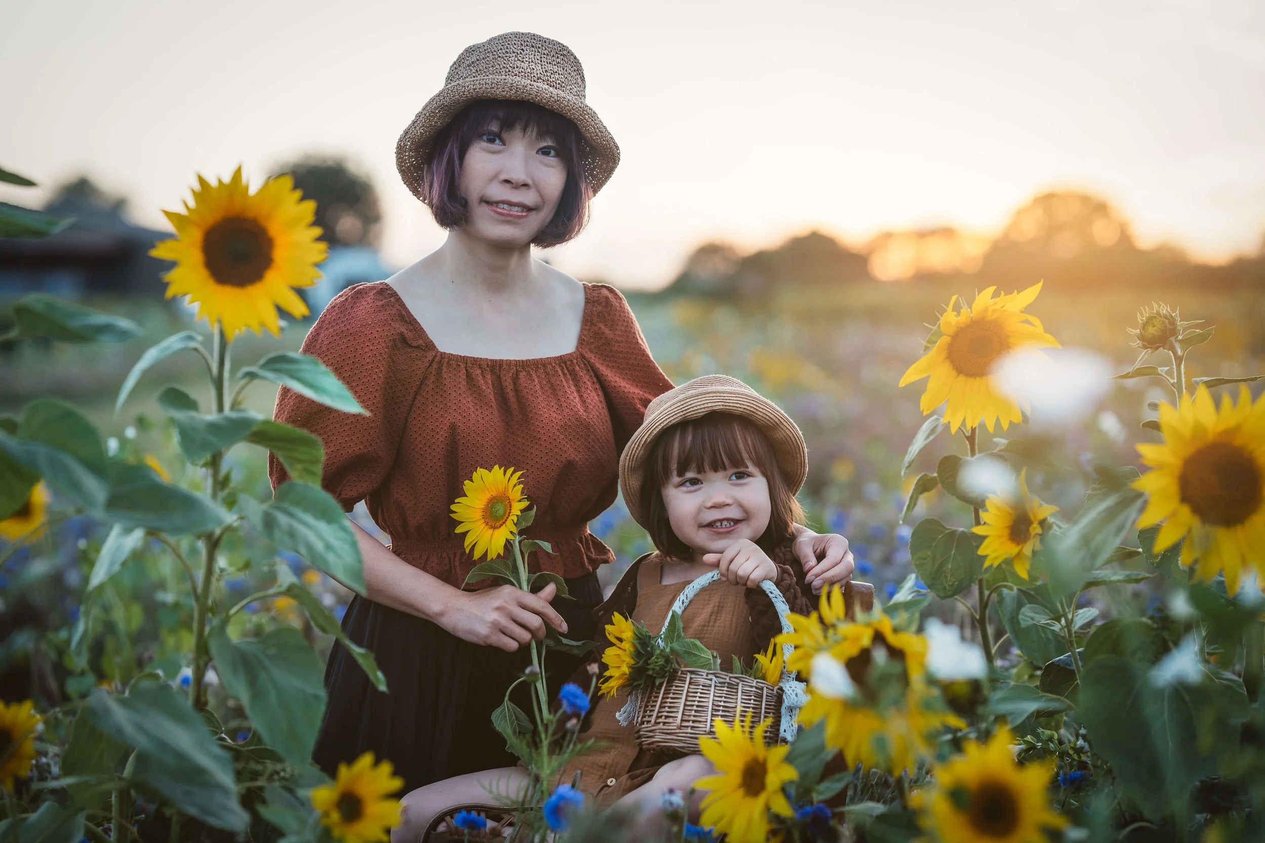 A woman and a young girl picking sunflowers in a field at sunset, with the woman holding a sunflower and the girl sitting with a wicker basket, both smiling.