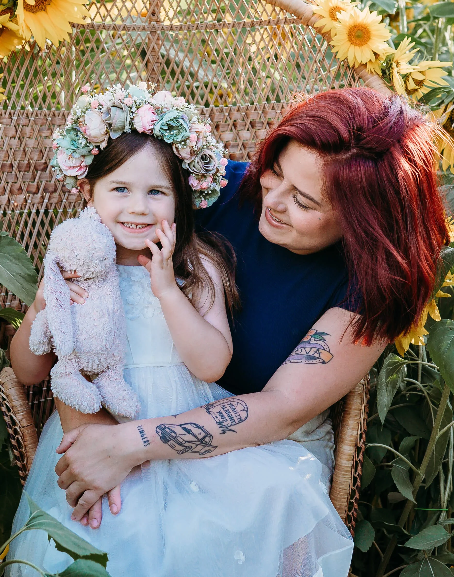 A woman with red hair and tattoos on her arms sitting on a wicker chair, holding a young girl with a floral crown and a light blue dress. The girl is smiling and holding a plush toy, surrounded by sunflowers and greenery.