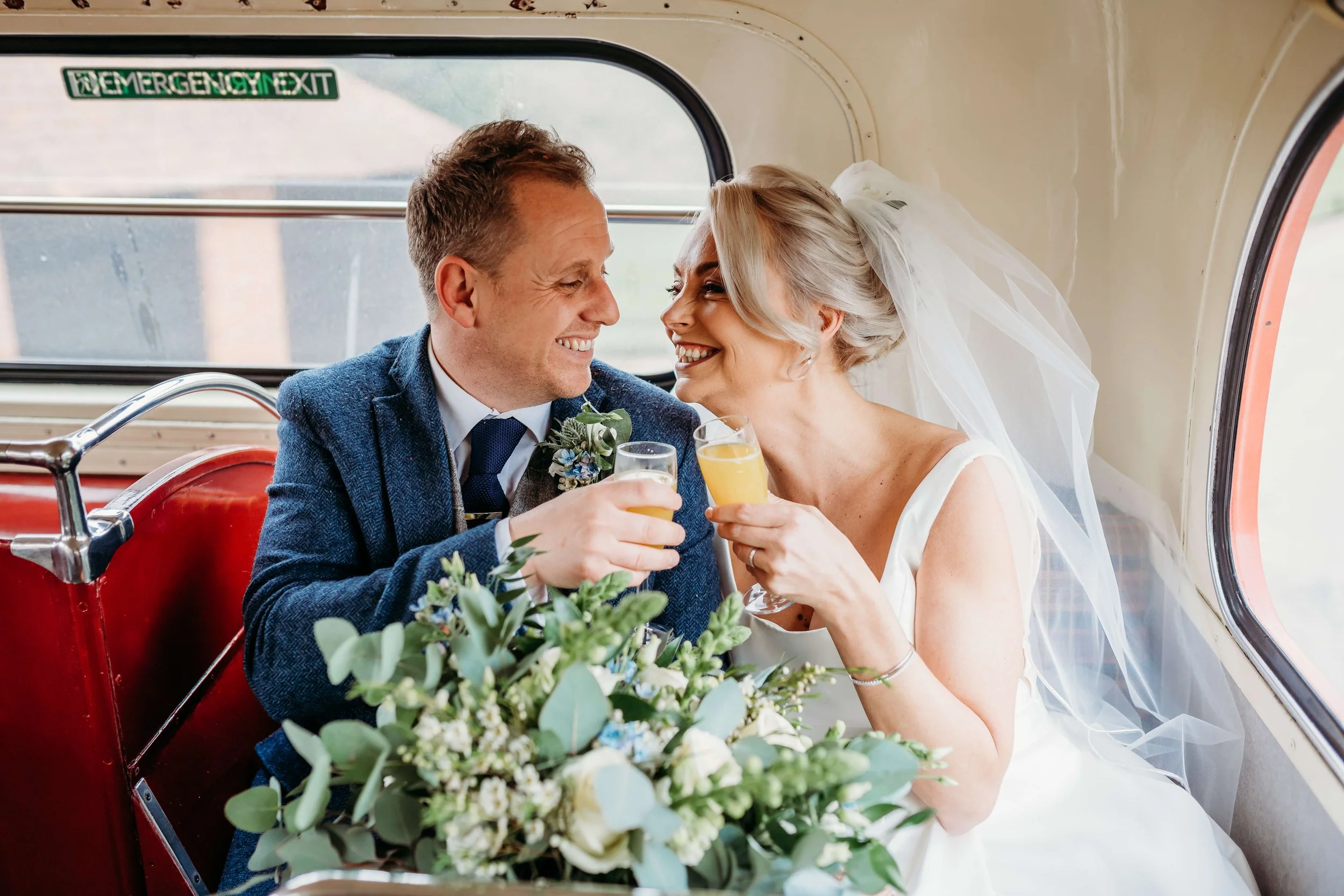 A newlywed couple smiling and toasting with drinks inside a vintage vehicle, with a bouquet of flowers in the foreground