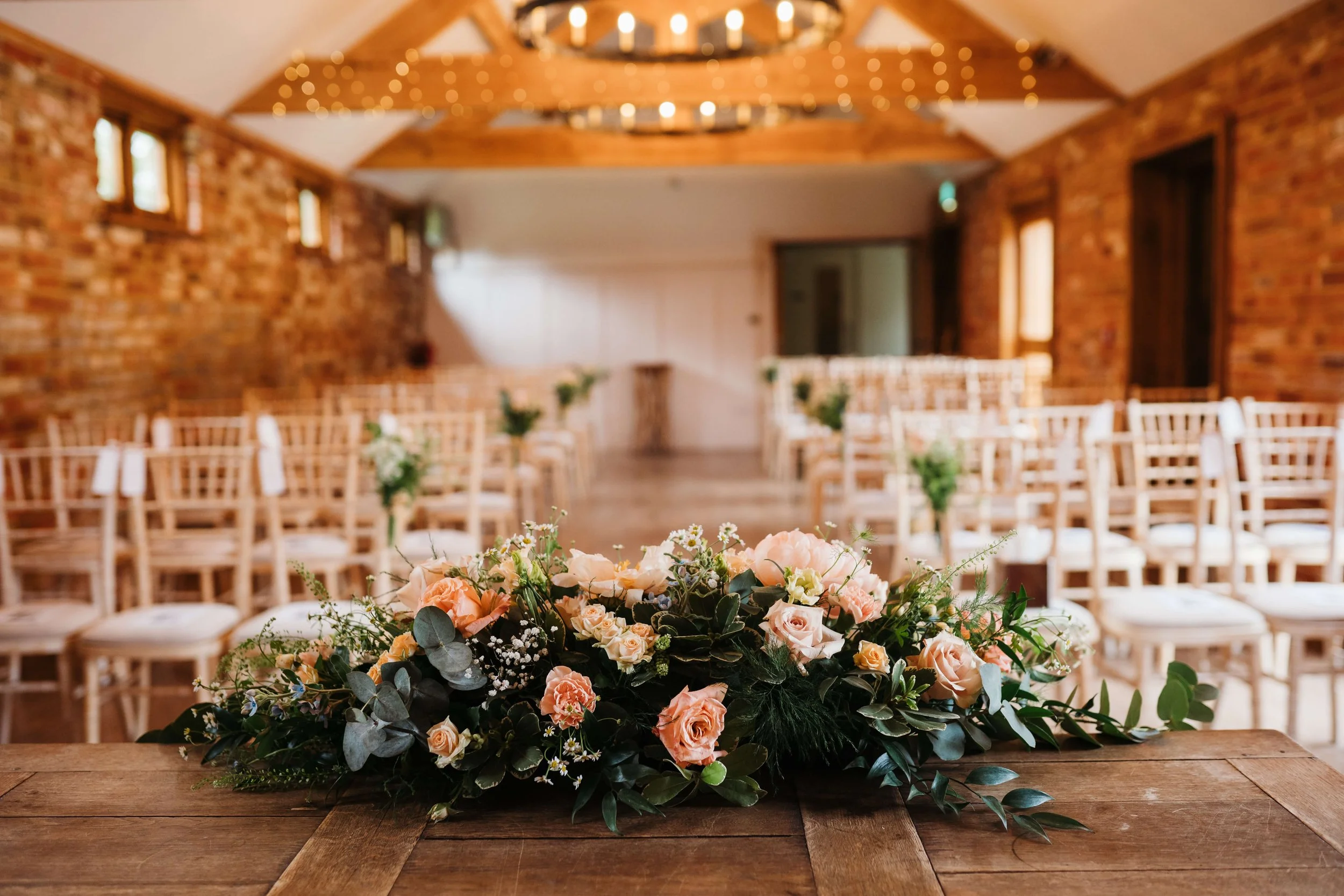 A floral arrangement with pink roses and greenery on a wooden table in an indoor wedding or event space with rows of chairs and brick walls.