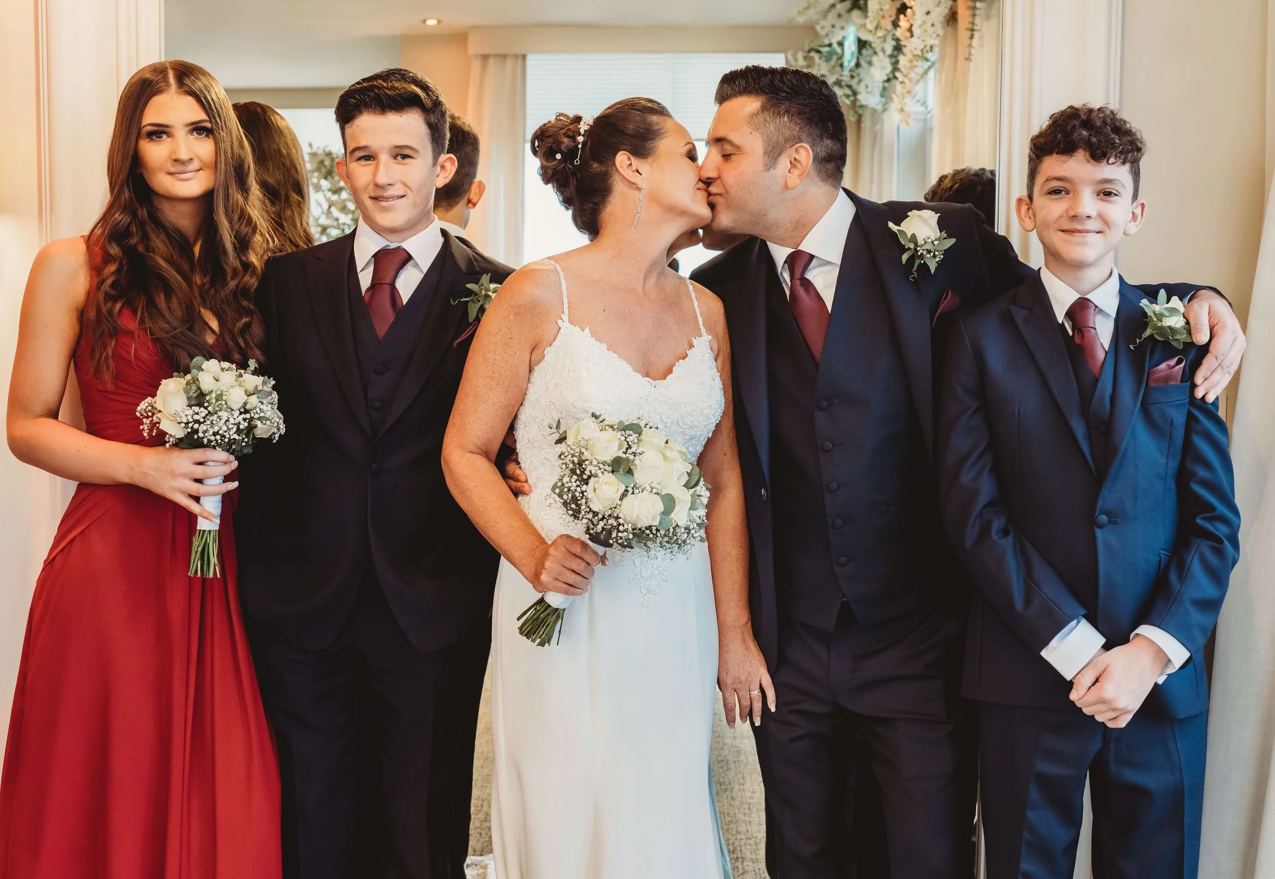 A wedding party consisting of a bride and groom kissing, two young men, and a young woman, all dressed formally. The bride wears a white gown and holds a bouquet, the groom and two young men wear dark suits with ties, and the young woman wears a red 