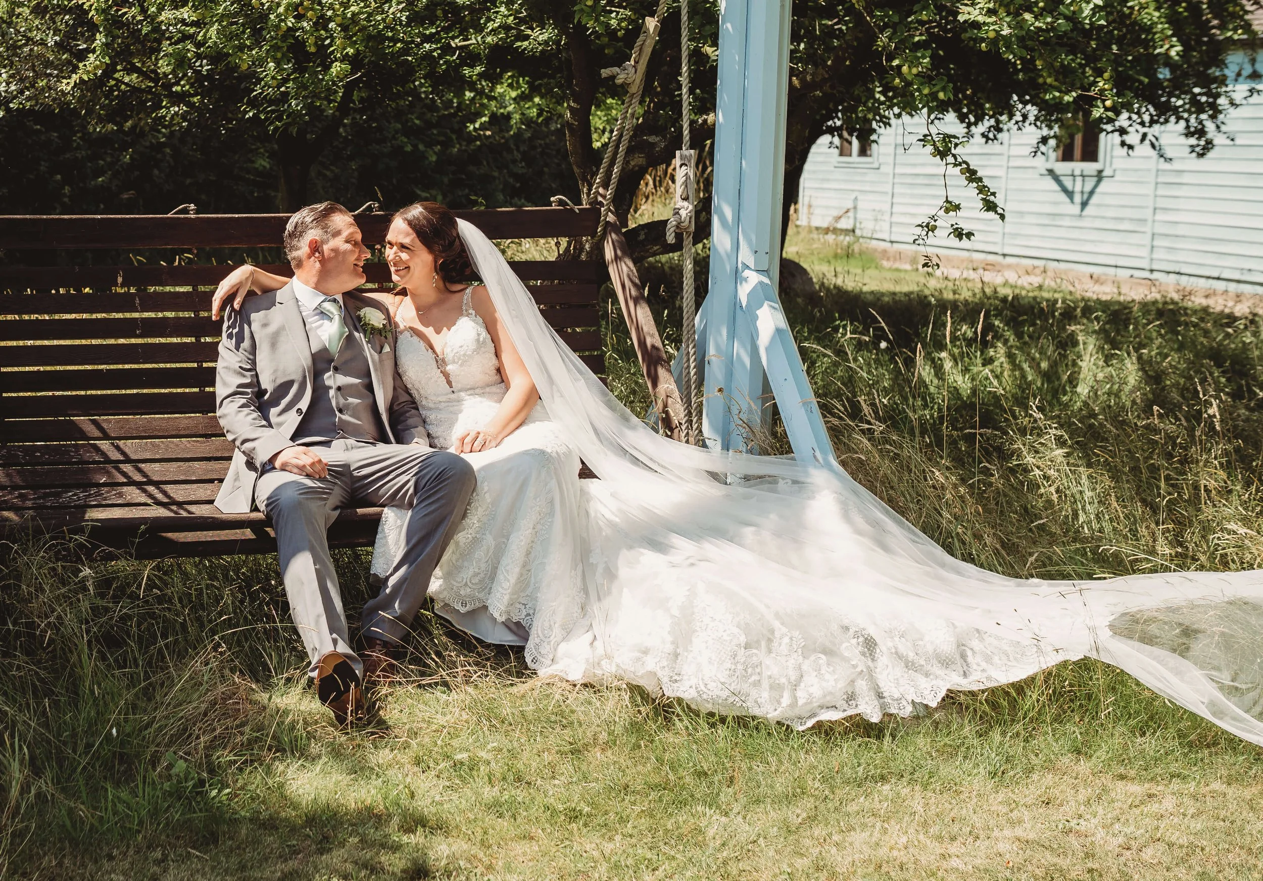 A bride and groom sitting on a wooden swing outdoors, smiling and looking at each other.