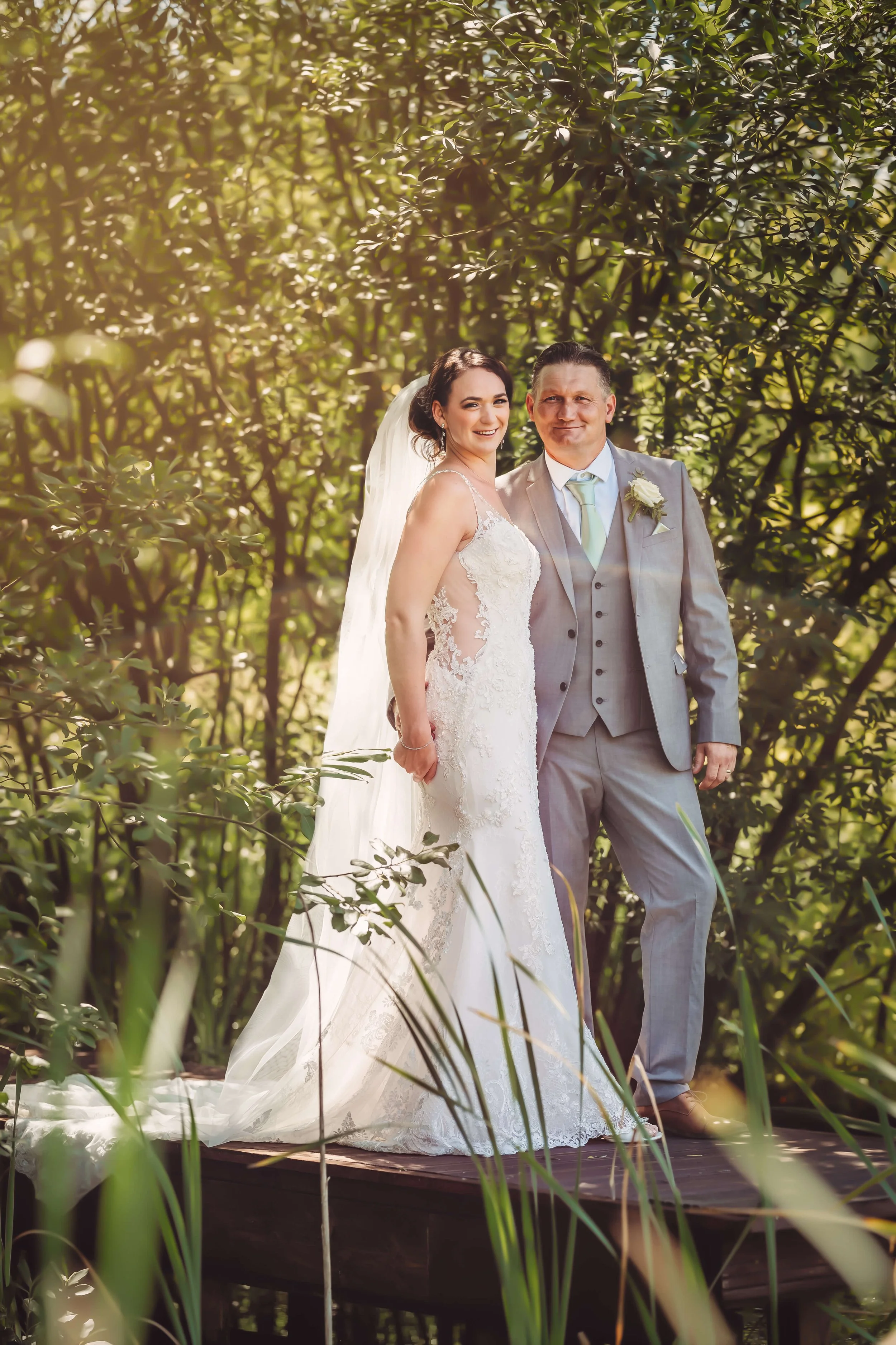Bride and groom standing on a wooden platform in a lush, green outdoor setting surrounded by trees, dressed in wedding attire, smiling at the camera.