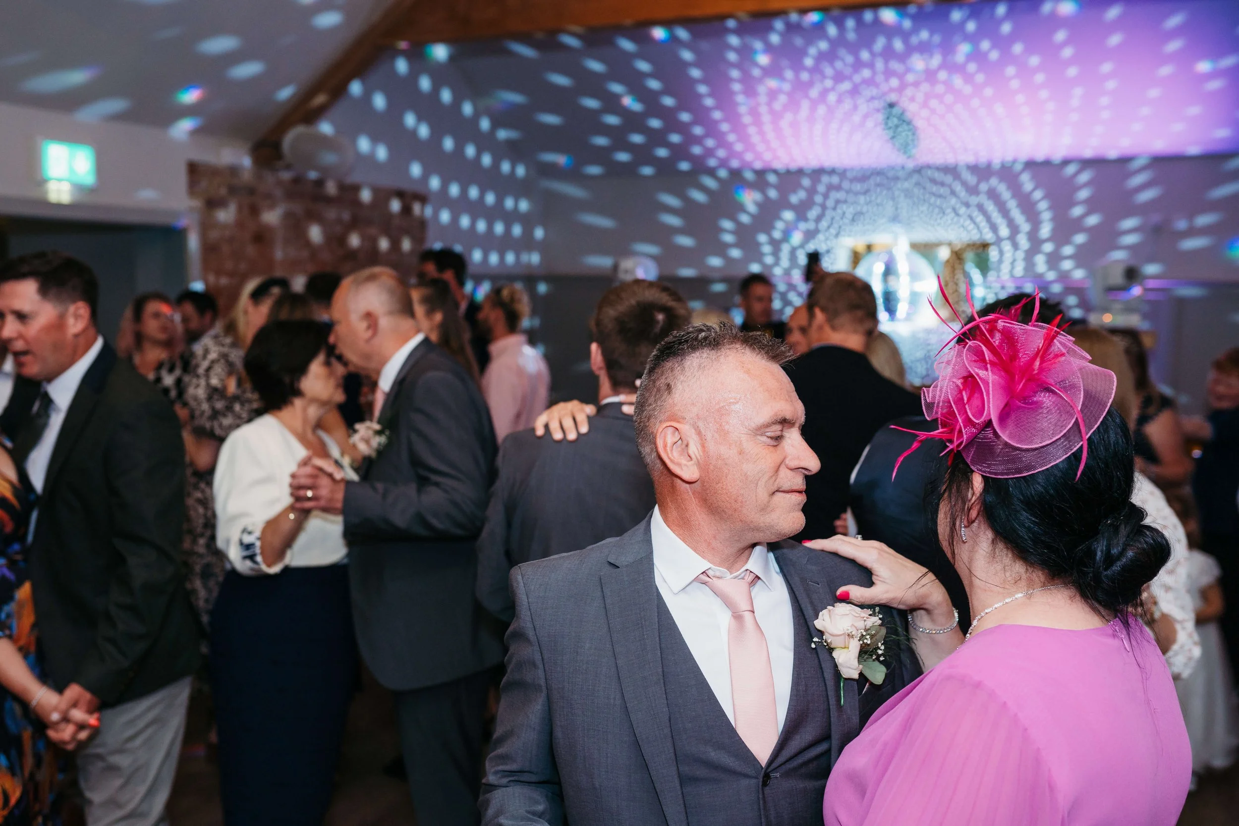 People dancing at a wedding reception, with a man and woman in the foreground sharing a dance, surrounded by guests in formal attire, under colorful disco lights.