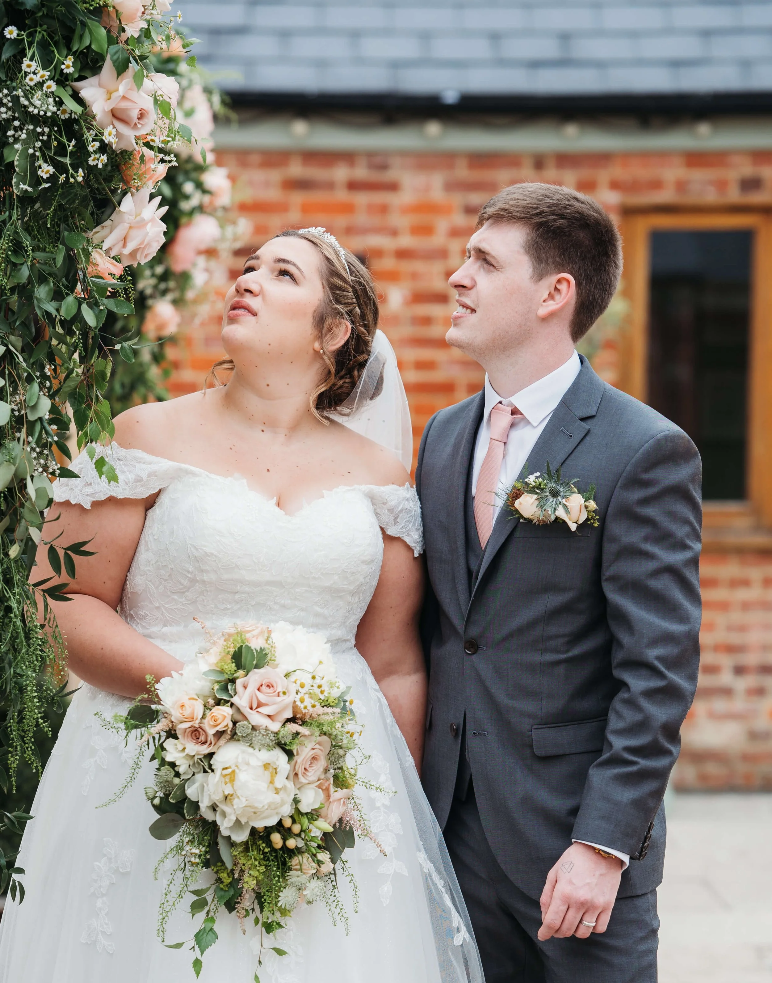 A bride and groom stand outdoors in front of a brick wall, with the bride holding a bouquet of flowers, looking up at a floral display or arch.