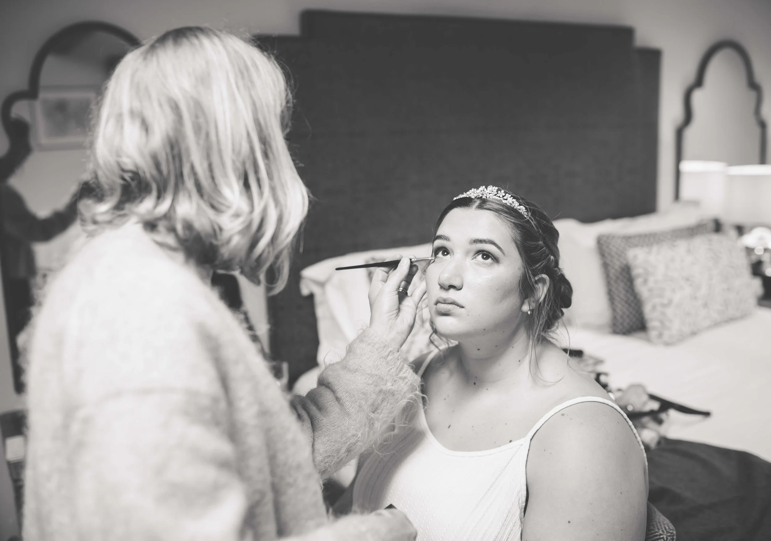 A woman with a tiara having her makeup done by a makeup artist in a hotel room.