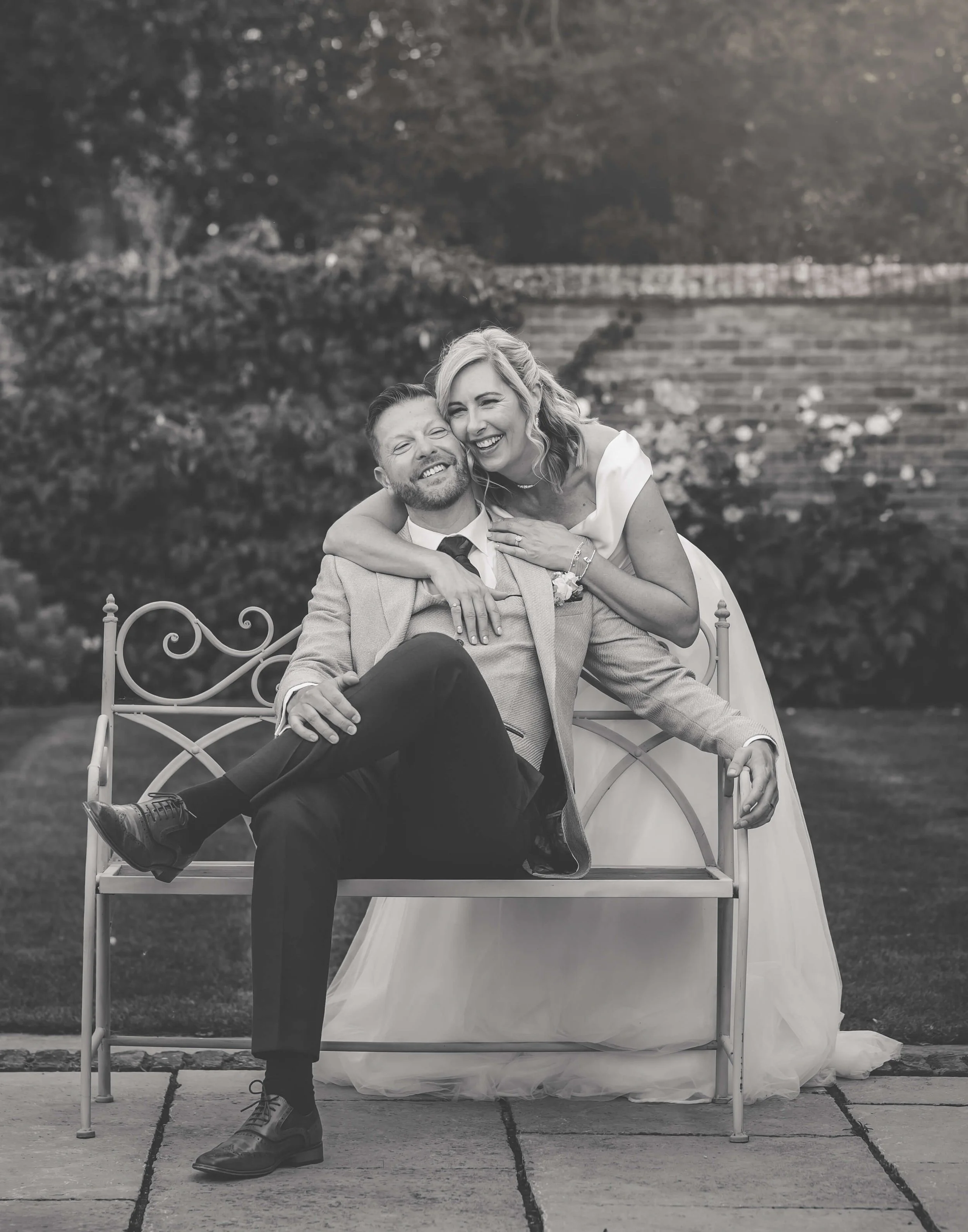 A black and white photo of a happy couple, with the woman in a wedding dress and the man in a suit, sitting on a metal bench outdoors, smiling and laughing together.