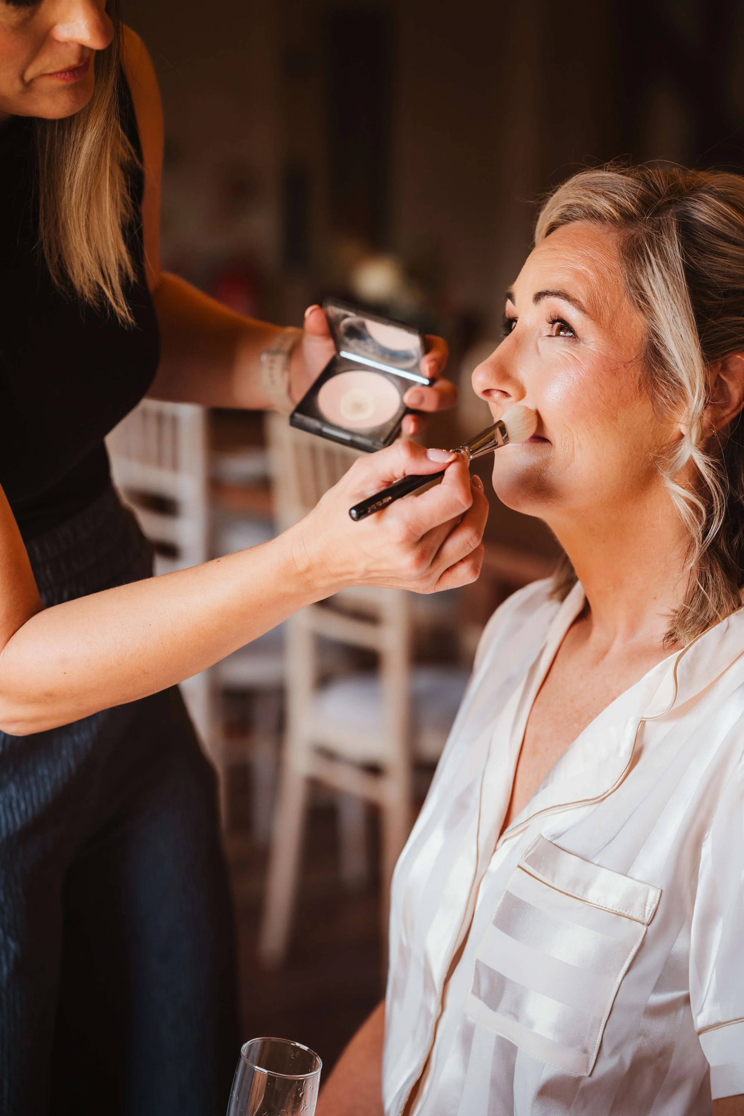A woman with styled hair and pajamas is having makeup applied by a makeup artist in a cozy room.