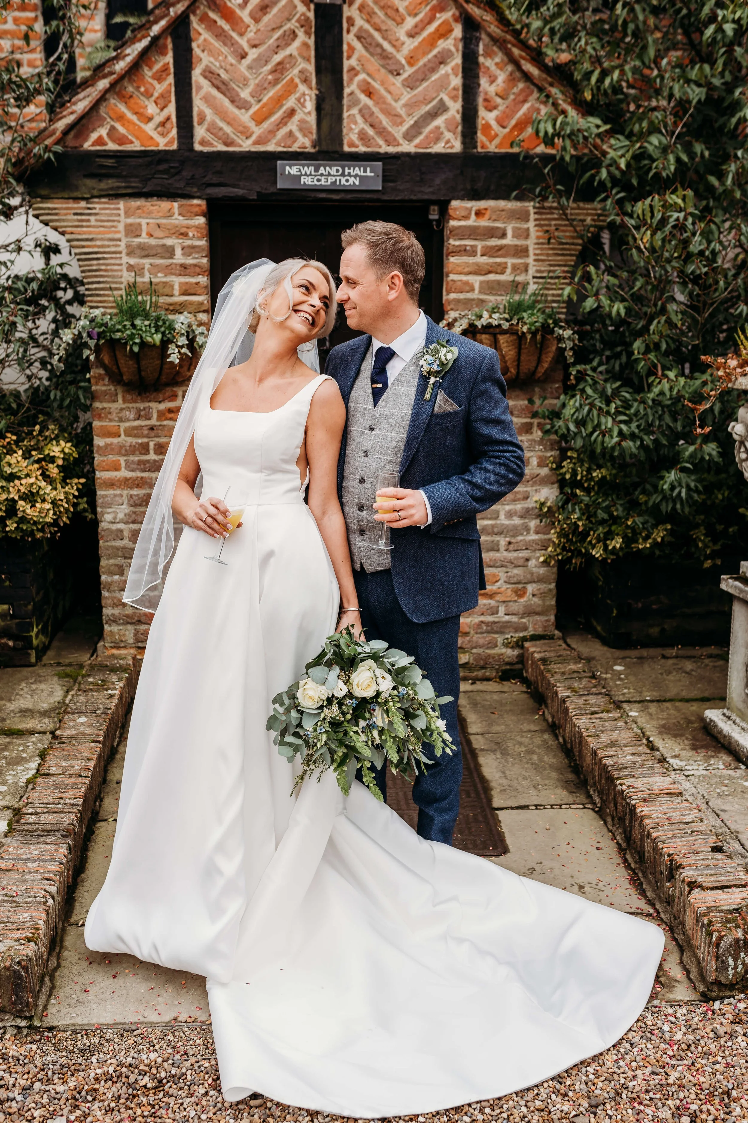 Bride and groom standing together outside a brick building, smiling and looking at each other, holding glasses of champagne. The bride is wearing a sleeveless white wedding gown with a veil, and holding a bouquet of white roses and greenery. The groo