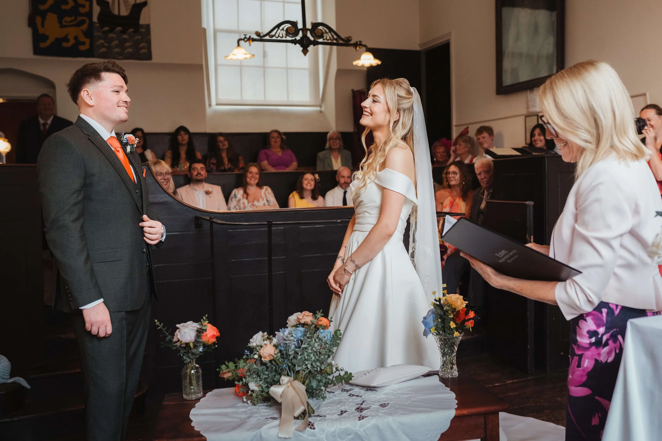 A wedding ceremony with a bride and groom facing each other, surrounded by guests in a church. The bride is in a white gown with off-the-shoulder sleeves, and the groom is in a gray suit with an orange tie. The officiant is reading from a book, and f