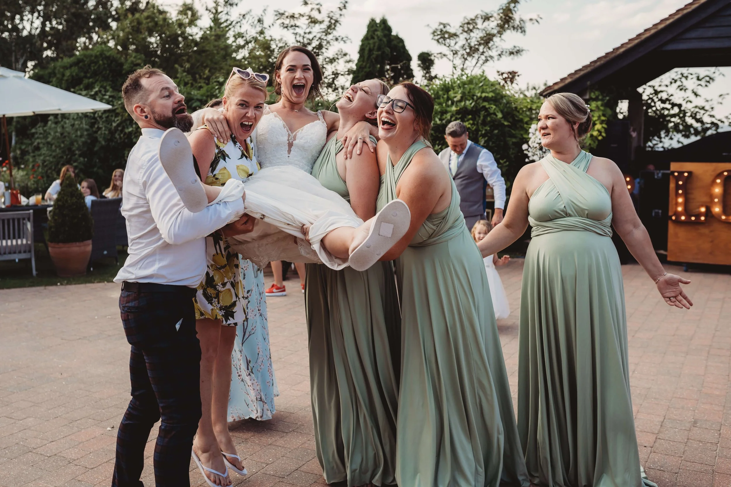 Group of people celebrating at a wedding, with women in green dresses and a man lifting a woman in a wedding dress, all smiling and laughing outdoors.