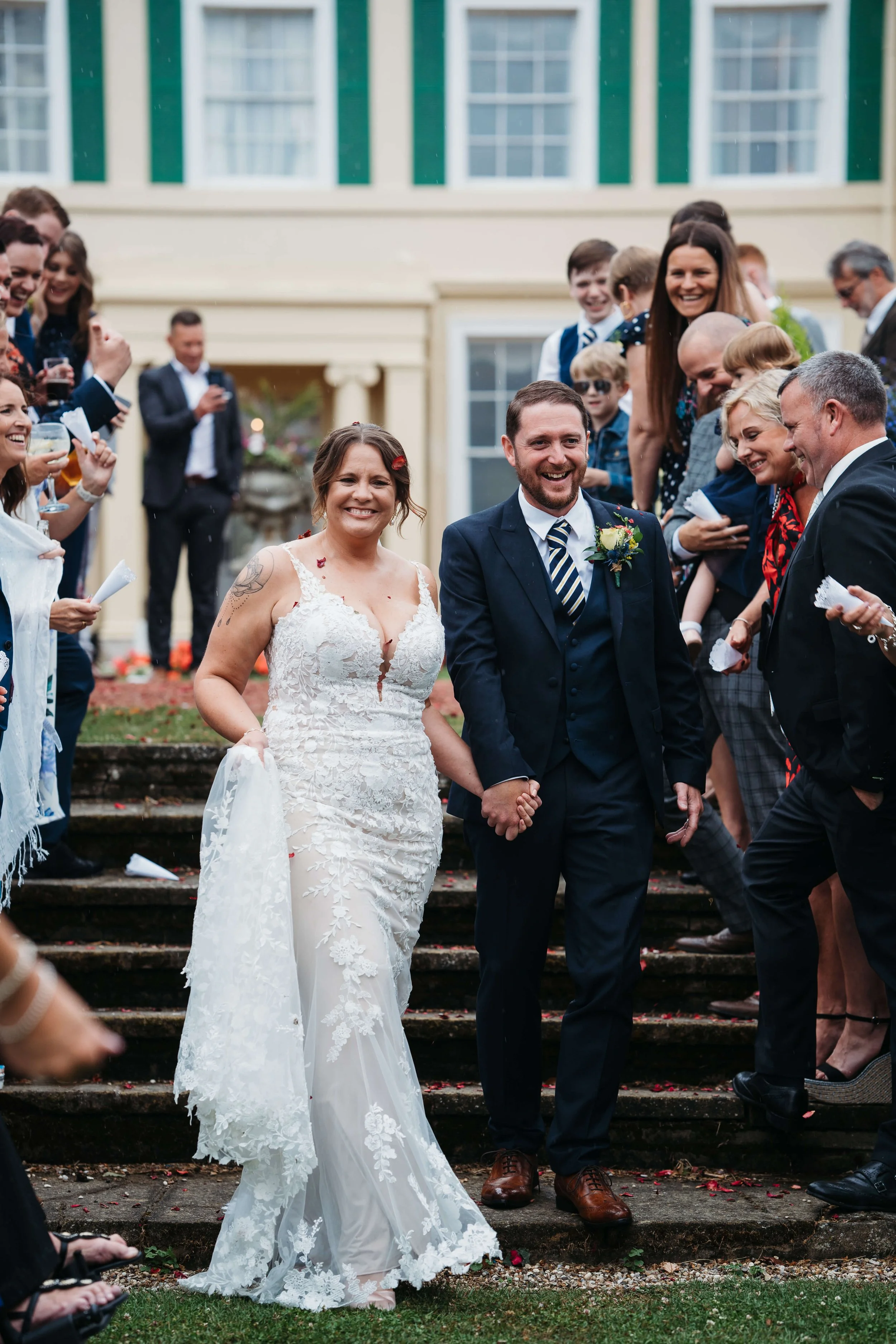 Bride and groom walking down the stairs, holding hands, smiling, surrounded by friends and family celebrating outdoors at a wedding.