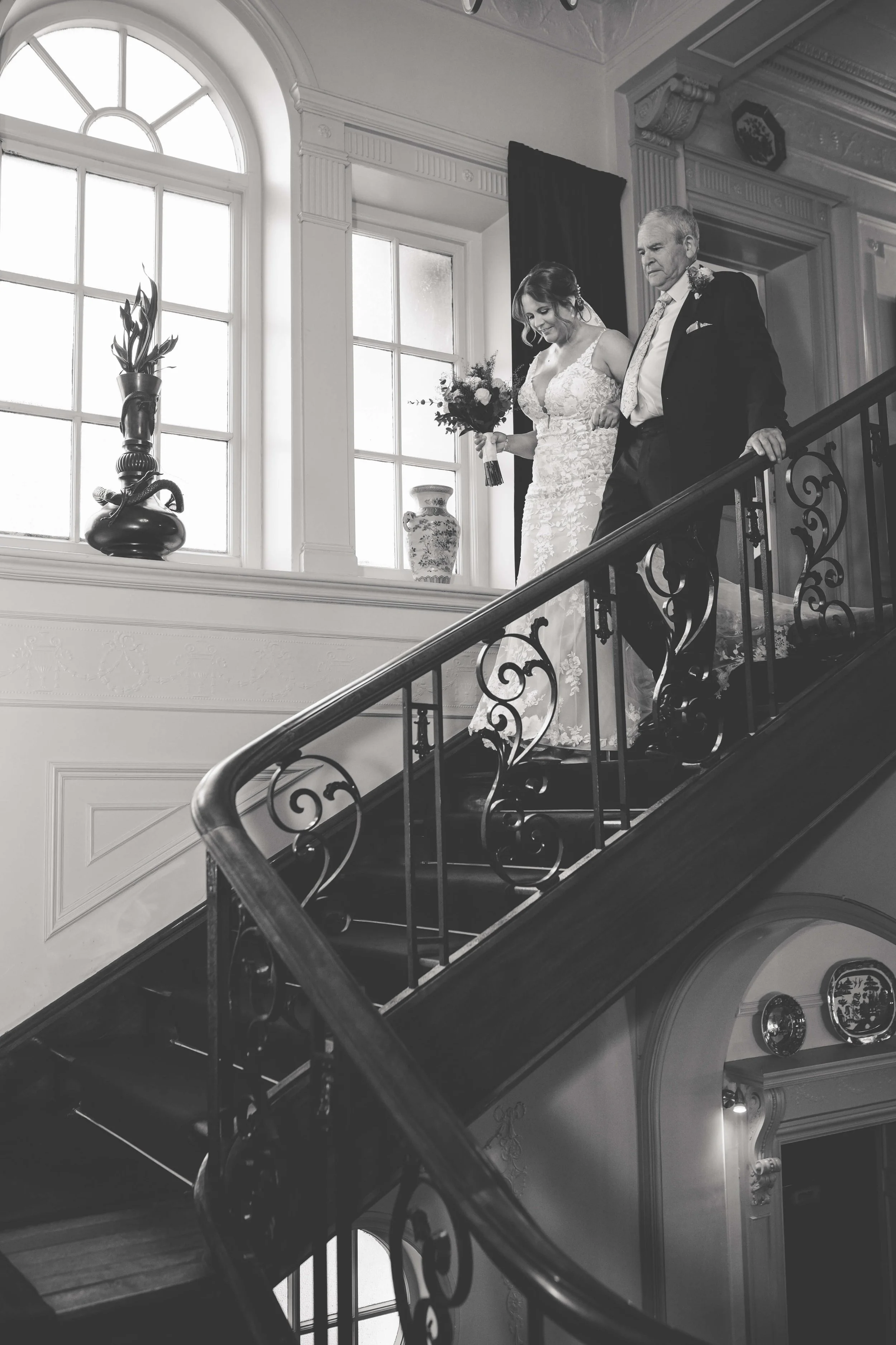 A bride and an older man, likely her father, descending a staircase inside a grand, well-lit building, possibly at a wedding.