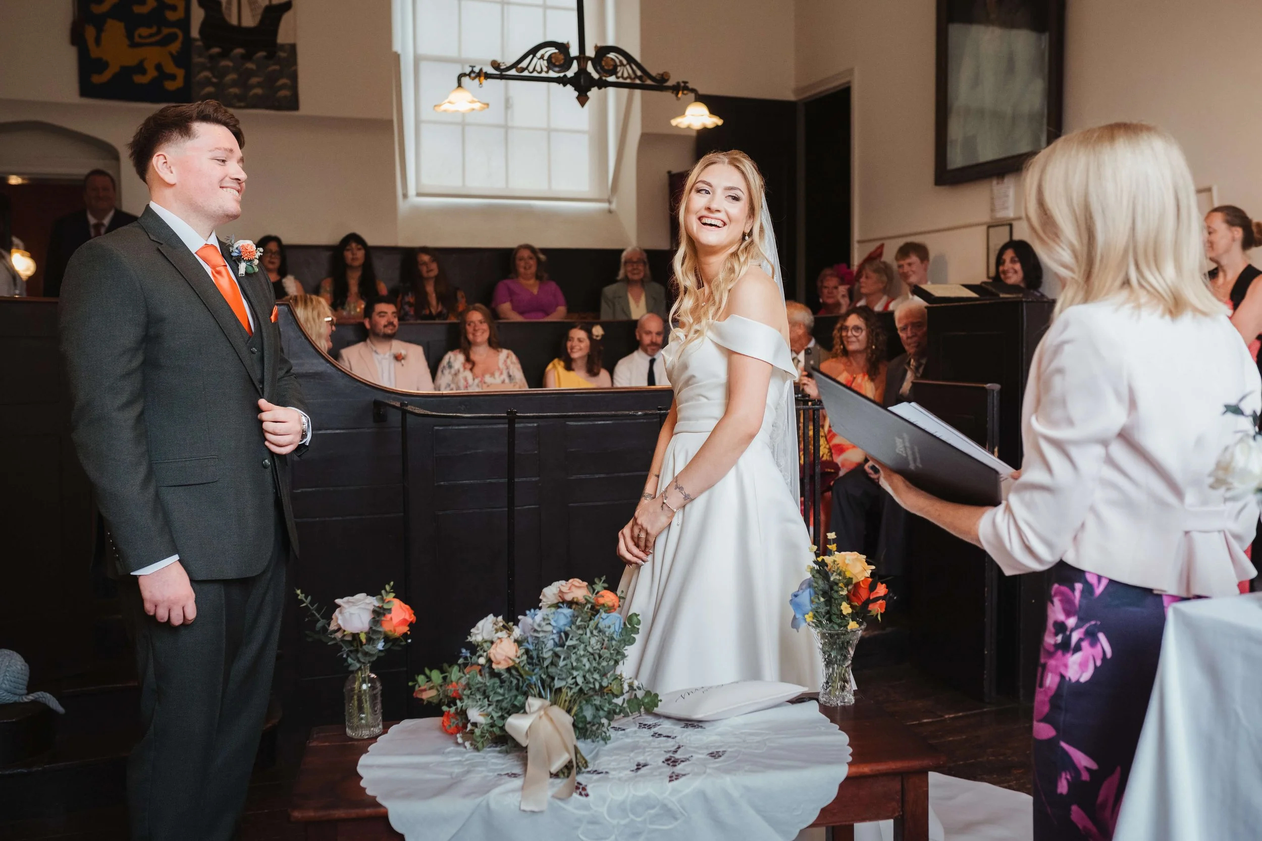 A bride and groom standing in front of a wedding officiant during their wedding ceremony, with guests seated and watching in the background.