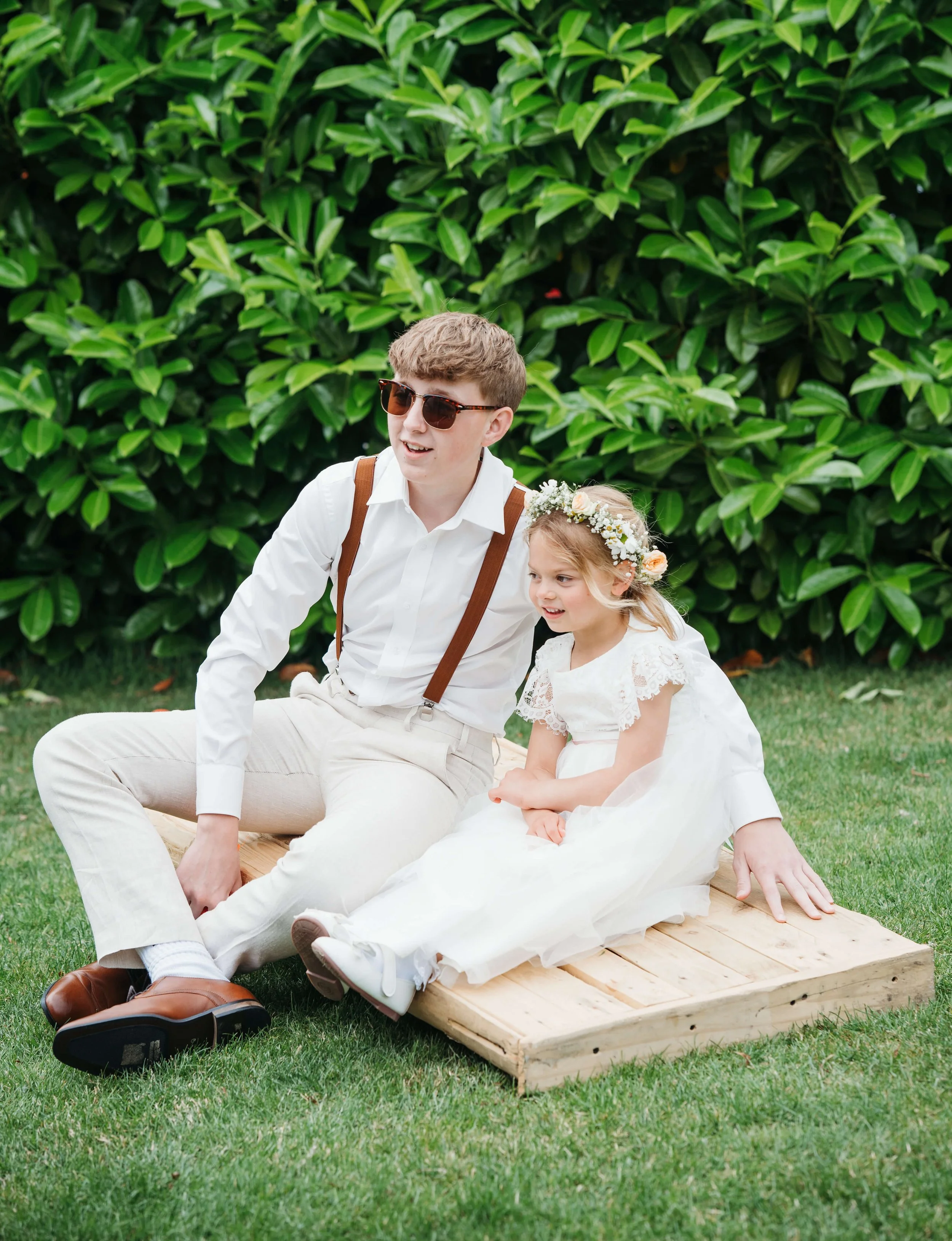 A young man and girl sitting on a wooden platform outdoors, with greenery in the background. The girl wears a white dress and floral headband, while the young man wears sunglasses, a white shirt, and suspenders.