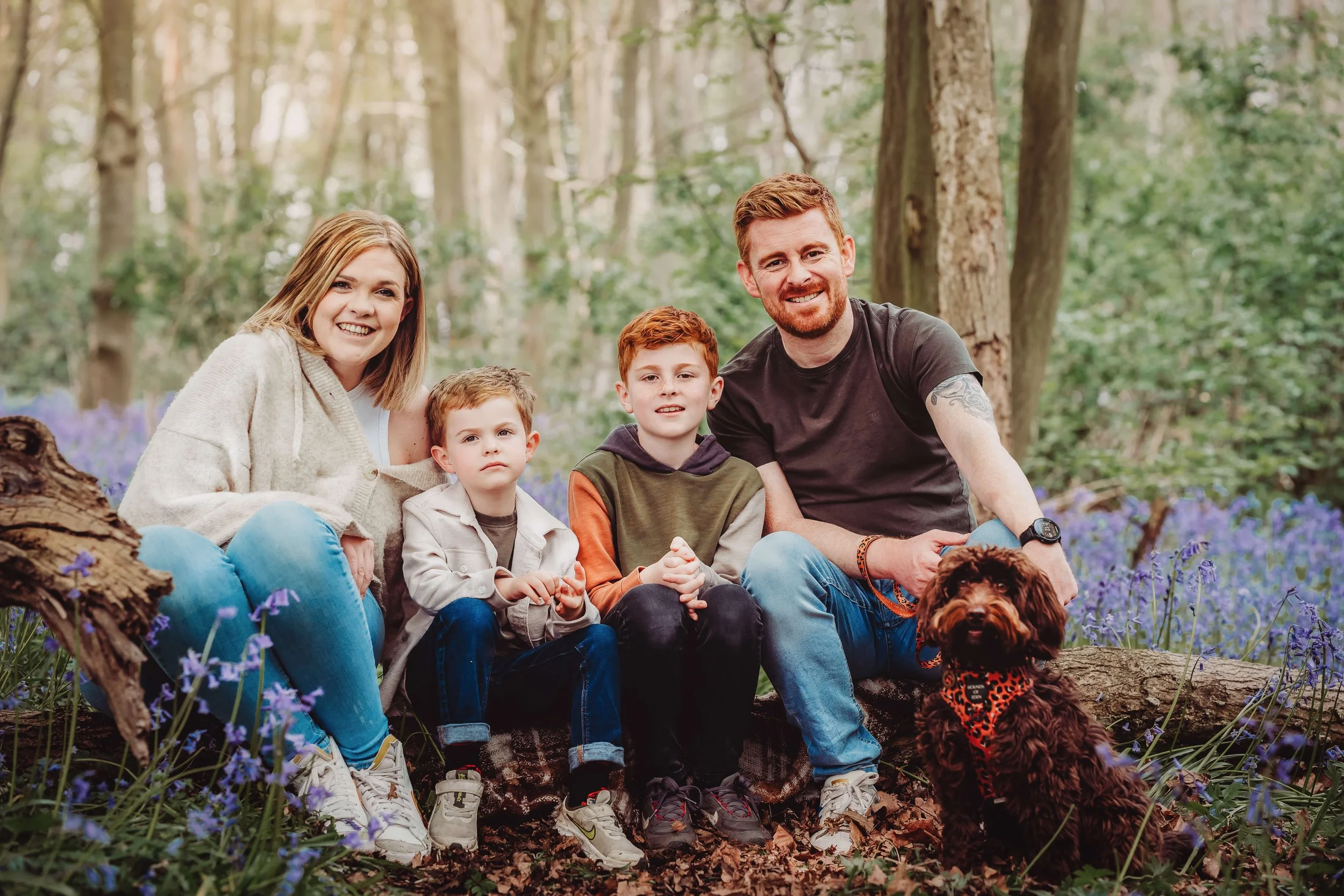 A family of five and their dog sitting on a fallen log in a wooded area surrounded by purple flowers, smiling at the camera.