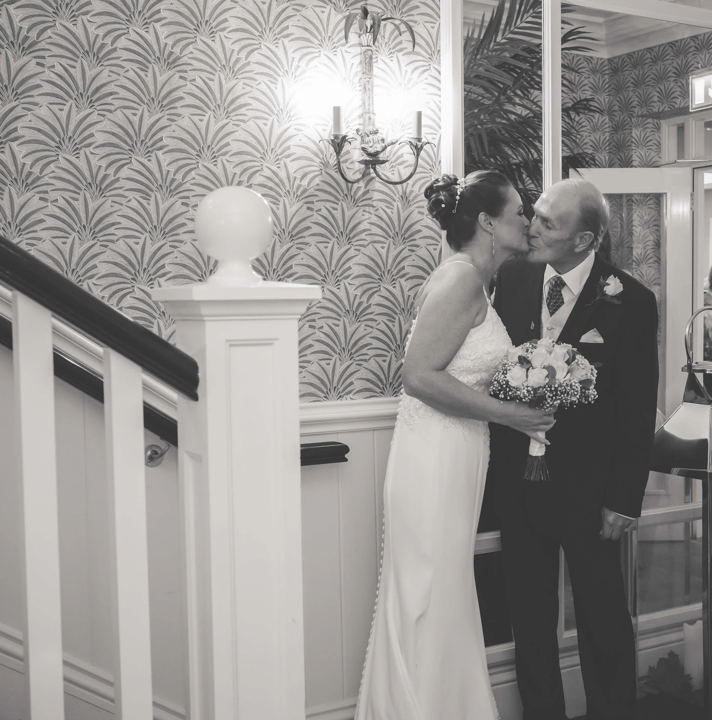 A black and white photo of a bride and an older man sharing a kiss in a decorated indoor venue. The bride is holding a bouquet and wearing a sleeveless white wedding gown, while the man is in a dark suit with a boutonniere.
